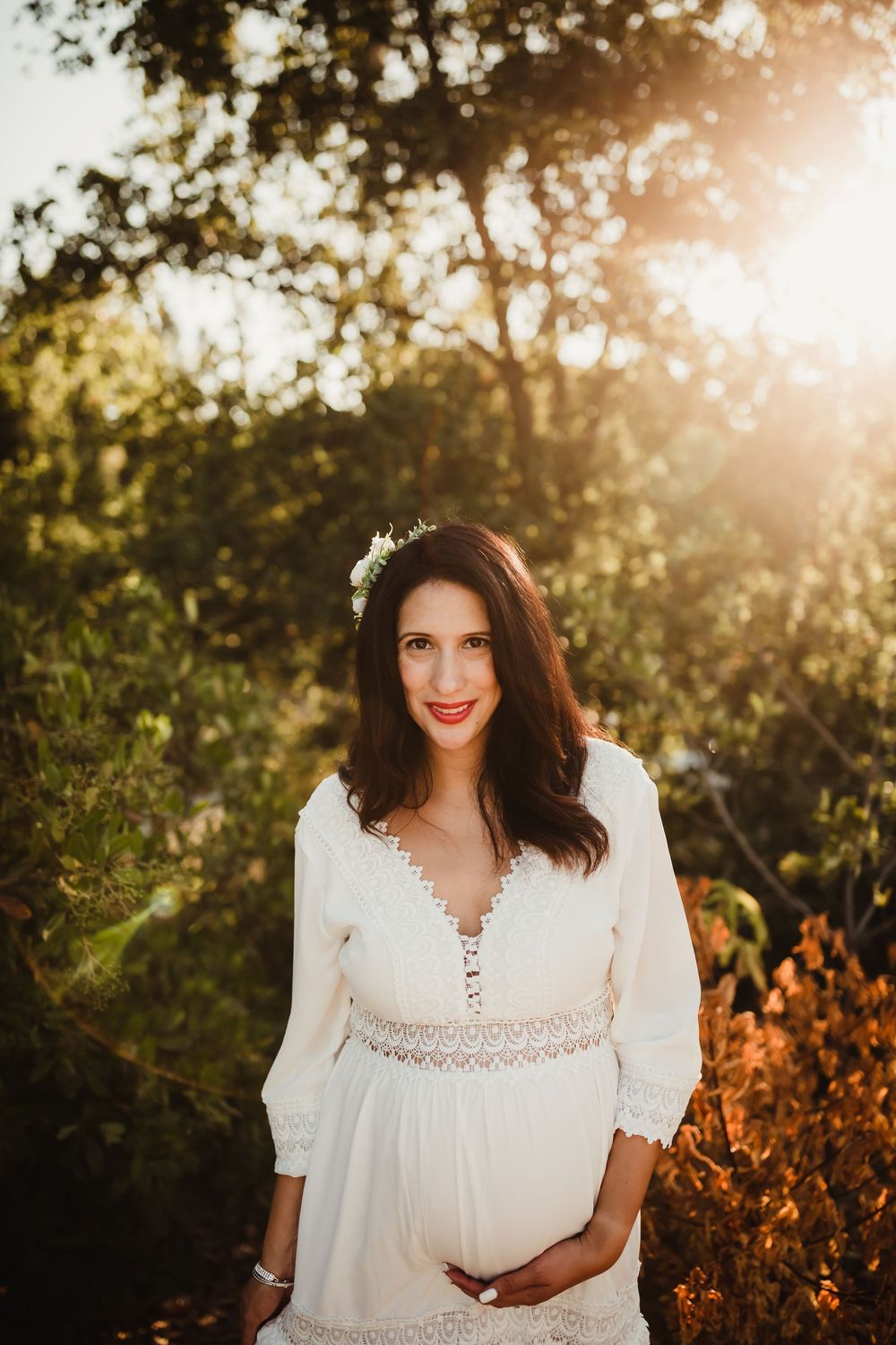 A woman in a white lace dress smiles in golden sunlight during an outdoor photoshoot in nature.