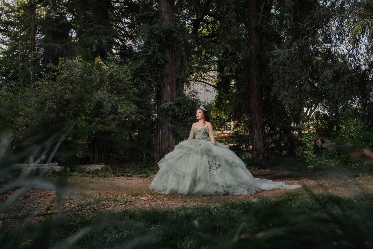 A bride in a flowing white wedding dress stands in a wooded forest setting during a photoshoot.