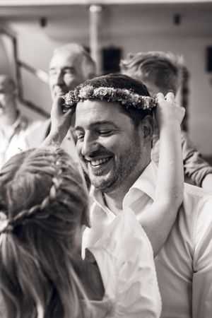 Black and white photo of a smiling man having a floral crown placed on his head at a wedding celebration.
