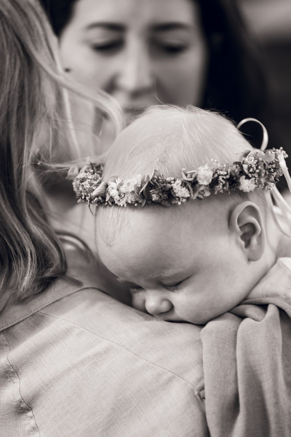 Black and white photo of a sleeping baby wearing a floral crown, cradled in a woman's arms.