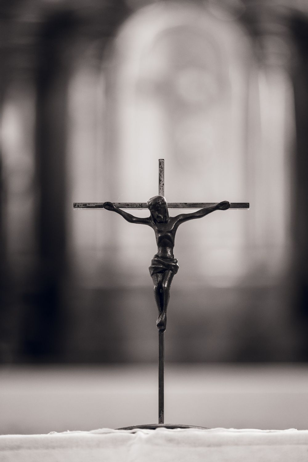 Black and white photo of a metal crucifix figurine on a stand, with a blurred church interior in the background.