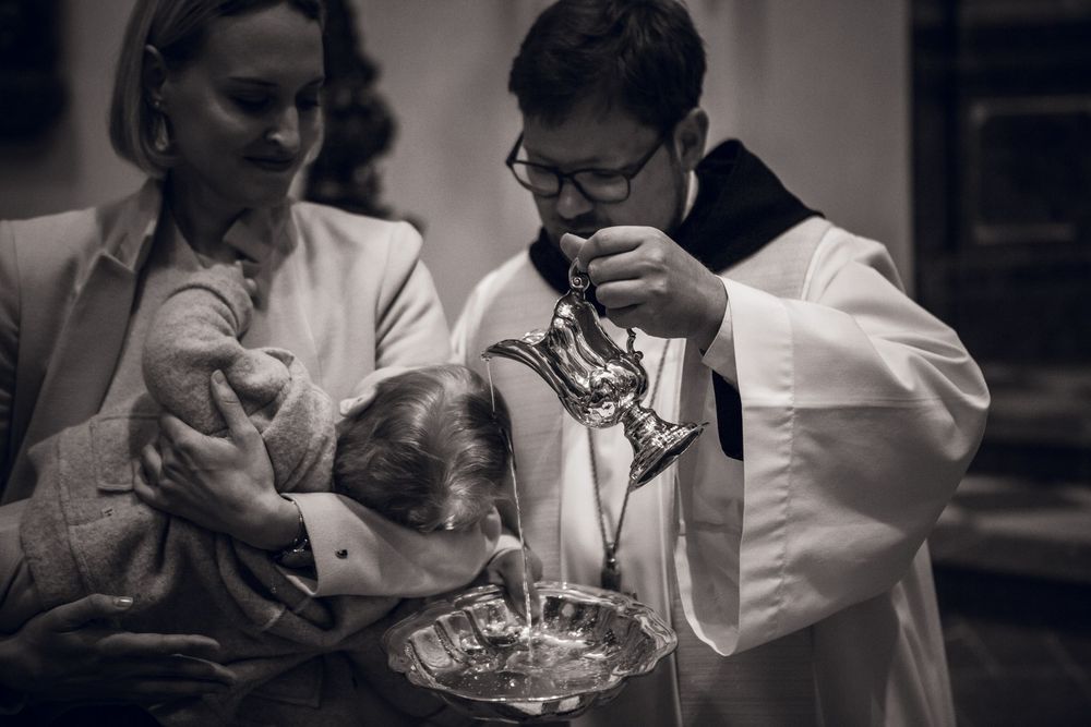 Black and white photo of a Catholic priest performing a baptism, pouring holy water over an infant held by a woman.