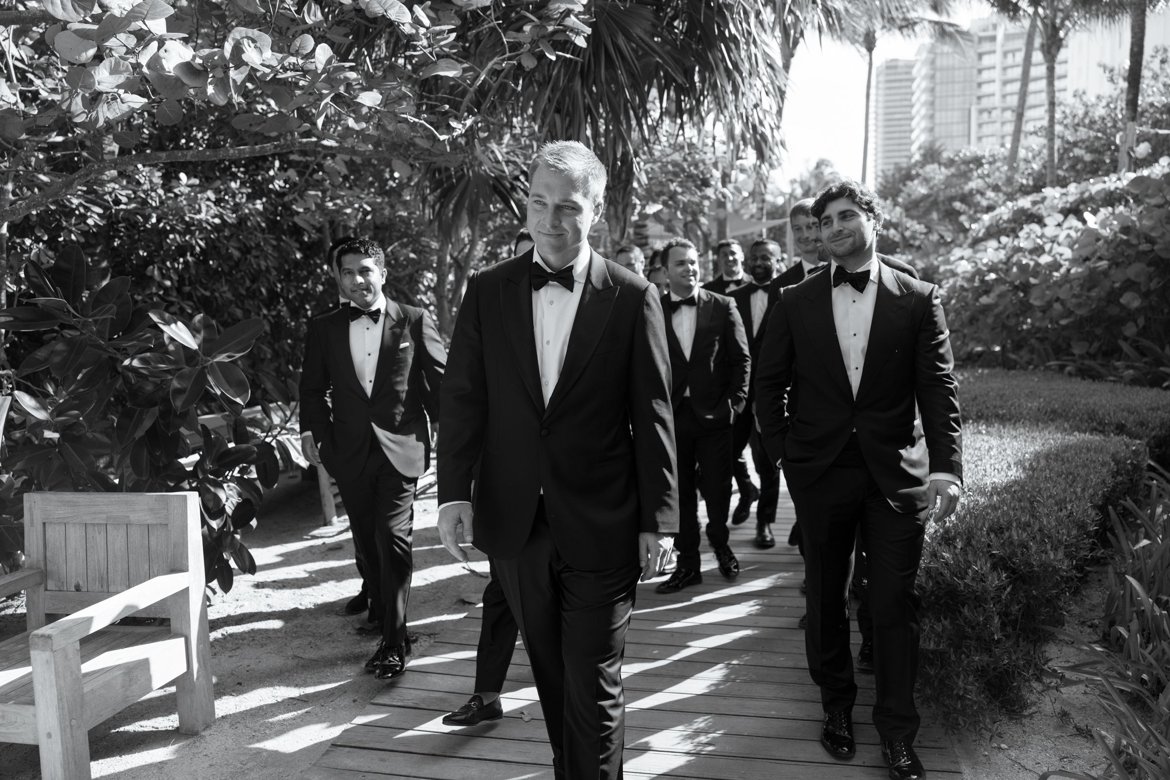 Groomsmen in black tuxedos and bow ties socialize during outdoor wedding reception beneath palm trees.