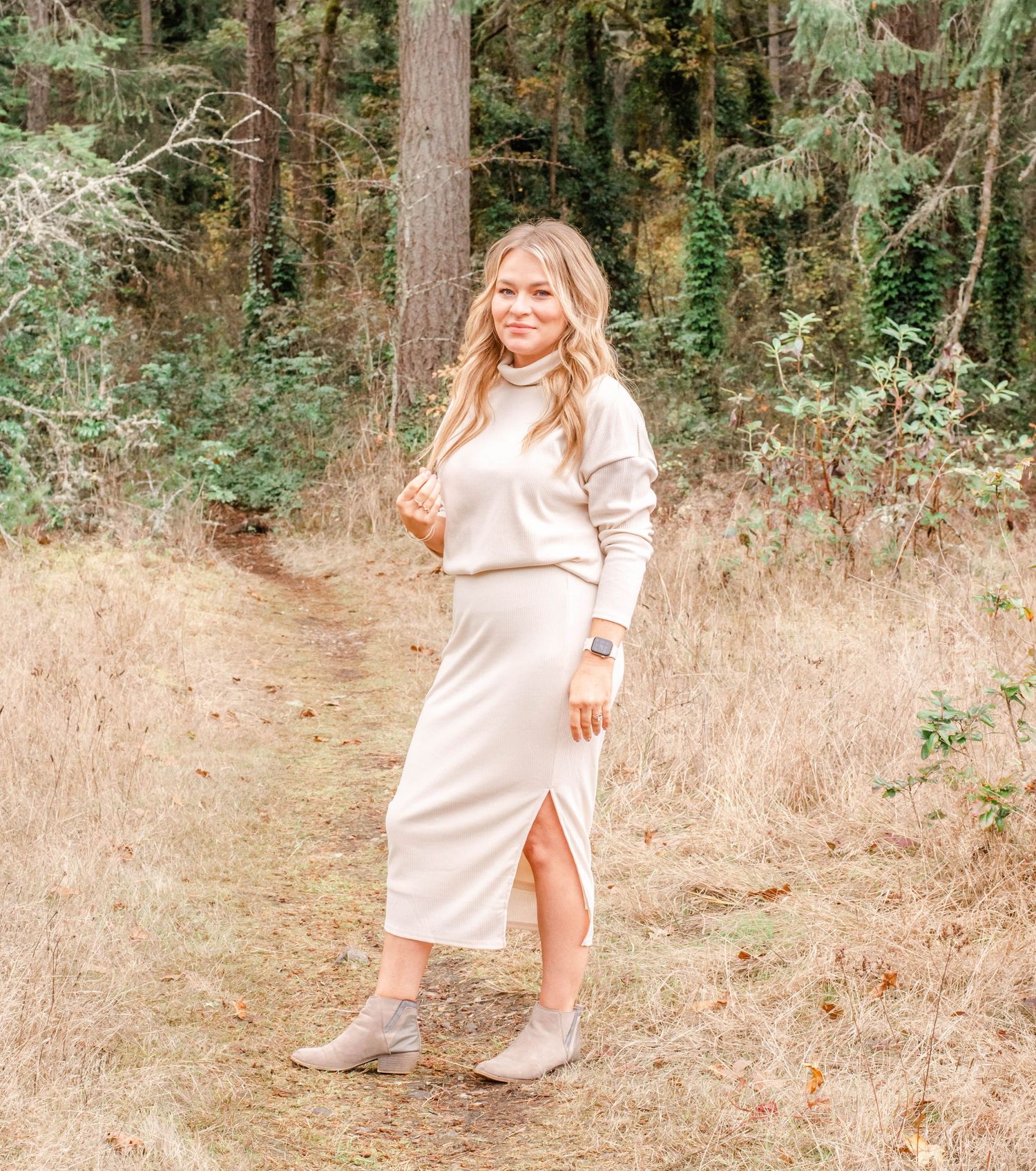 Person wearing cream-colored sweater dress poses in a forest setting with brown grass and green trees.