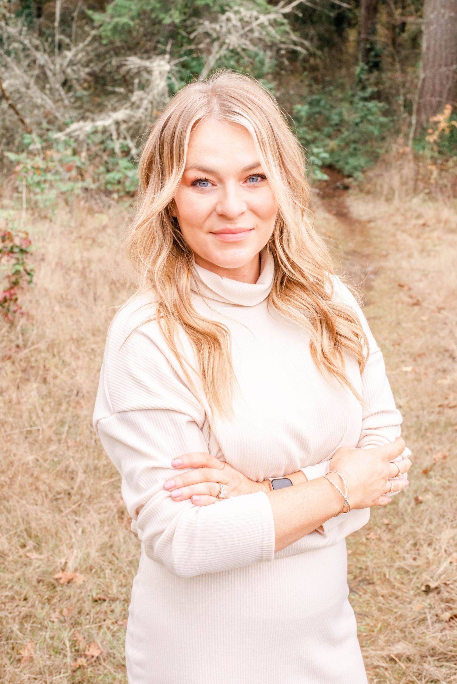 Individual in cream turtleneck poses with crossed arms against natural outdoor setting.
