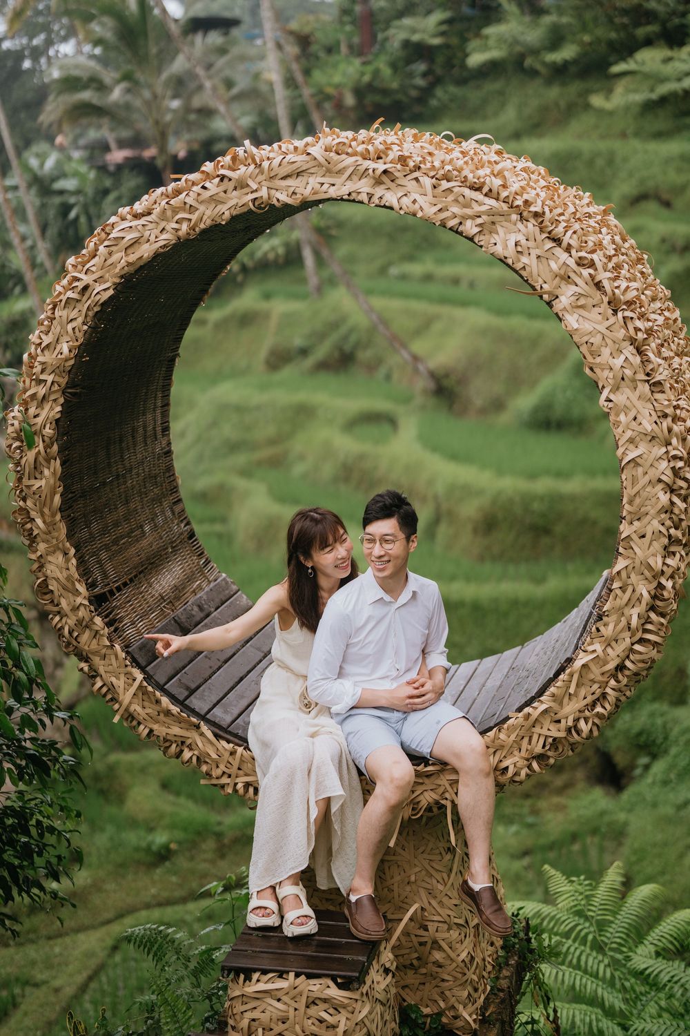 Two people sitting together on an ornate circular swing with tropical landscape behind them.