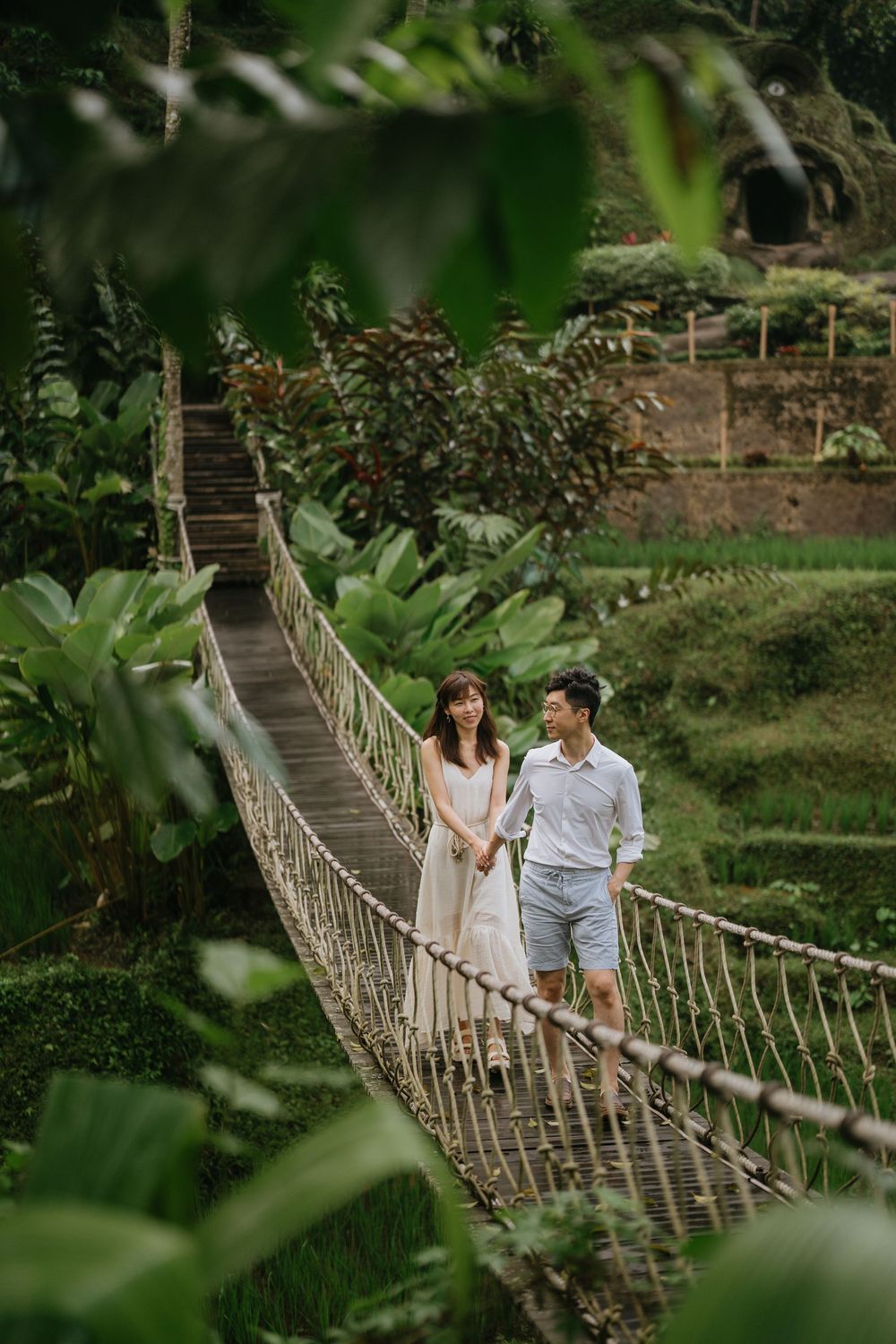 A couple walks across a rustic rope bridge surrounded by lush tropical foliage in a romantic outdoor setting.