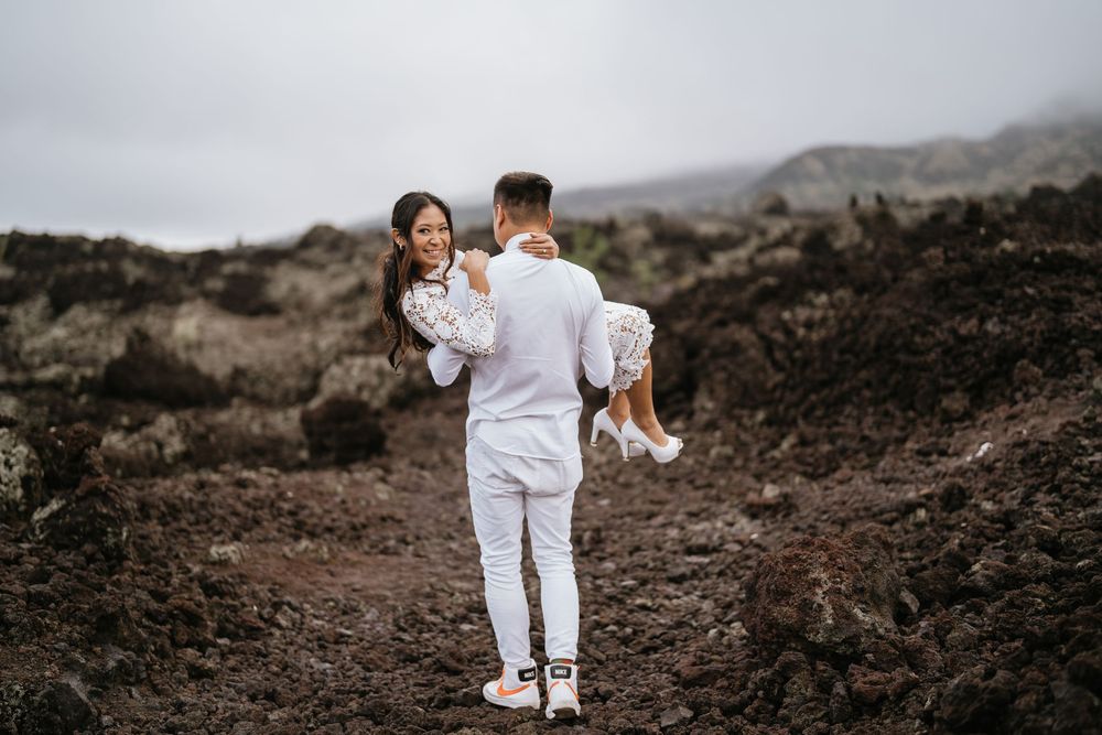 A couple in white outfits walk along a dramatic volcanic landscape with mountains in the background.