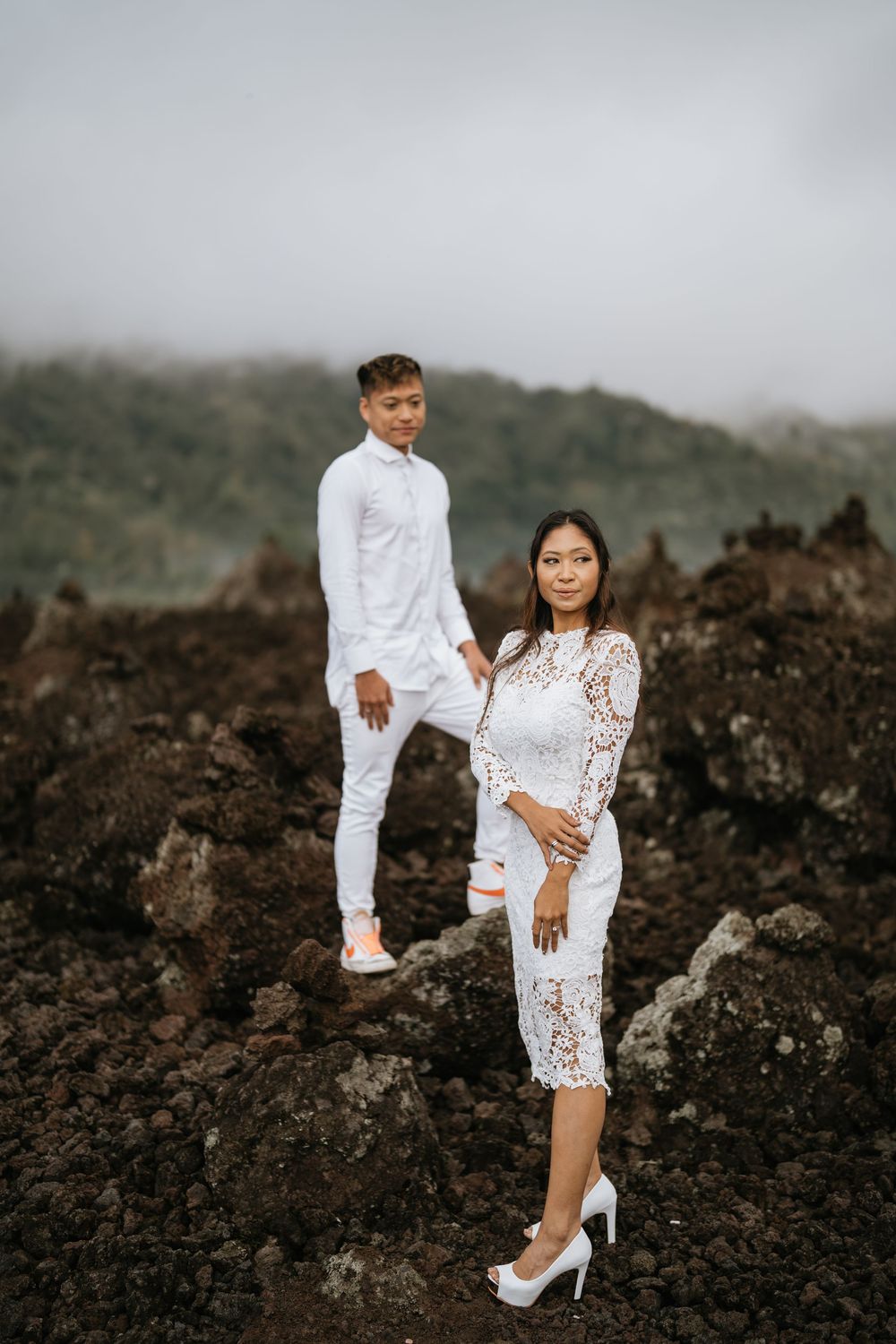 Couple in white outfits posing on volcanic rocks with misty mountains in background.