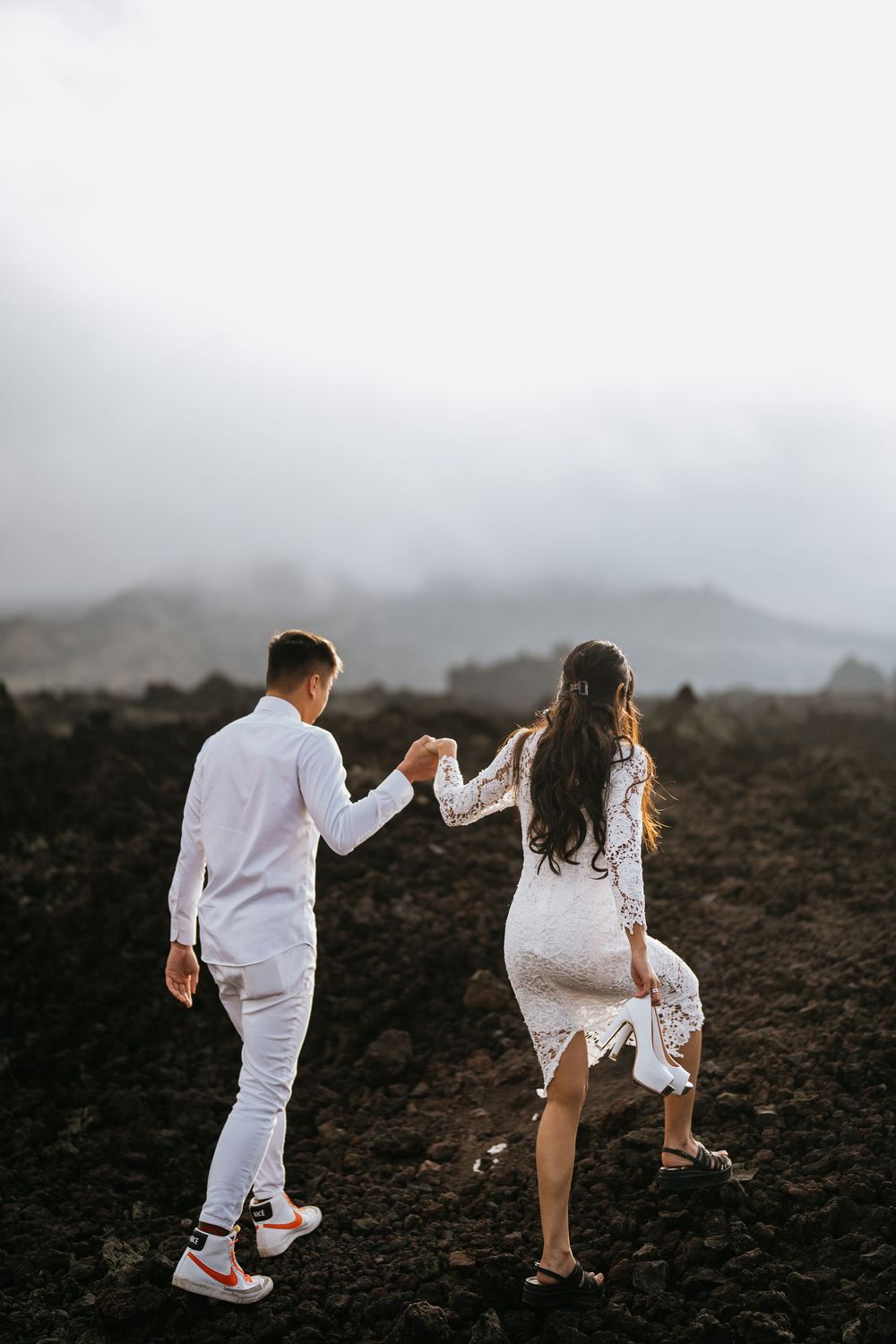Couple in white attire exploring rocky volcanic terrain against cloudy mountain backdrop.