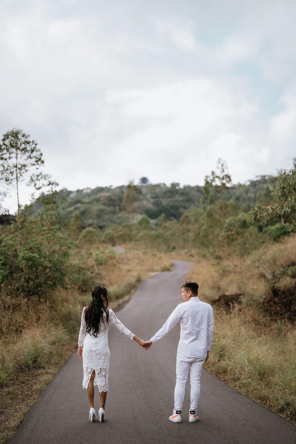 A couple in white outfits holding hands while walking down a winding mountain road.