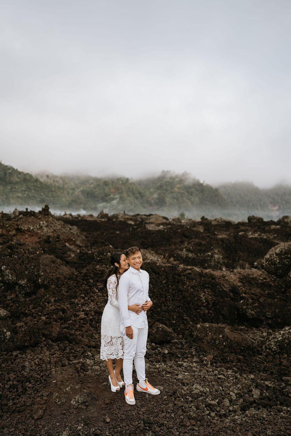 A couple dressed in white embracing on dark volcanic terrain with misty mountains in background.