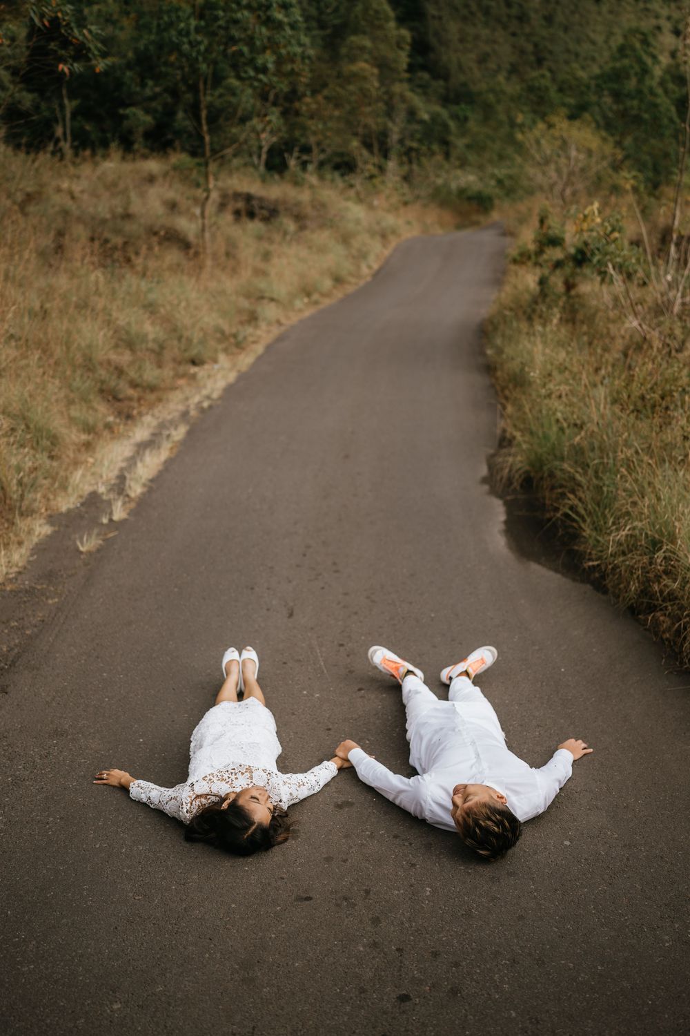 Two people in white outfits lying playfully on an asphalt road surrounded by greenery.