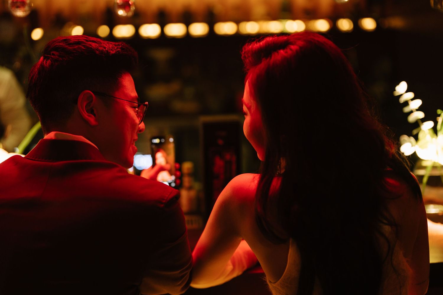 Intimate couple conversing closely at a bar bathed in warm red ambient lighting.