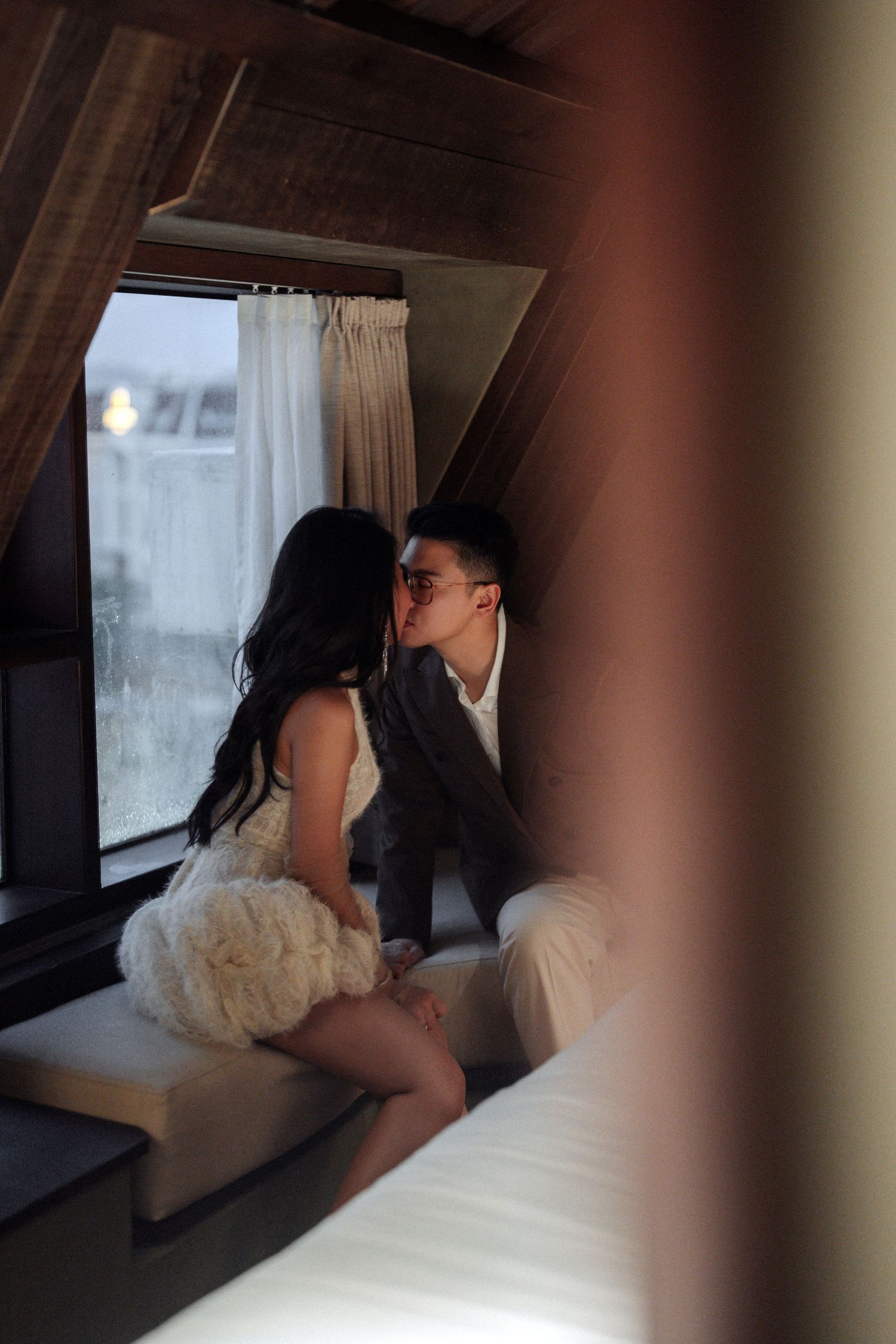 Couple sharing a kiss by attic window, bride in white feathered dress and groom in gray suit.