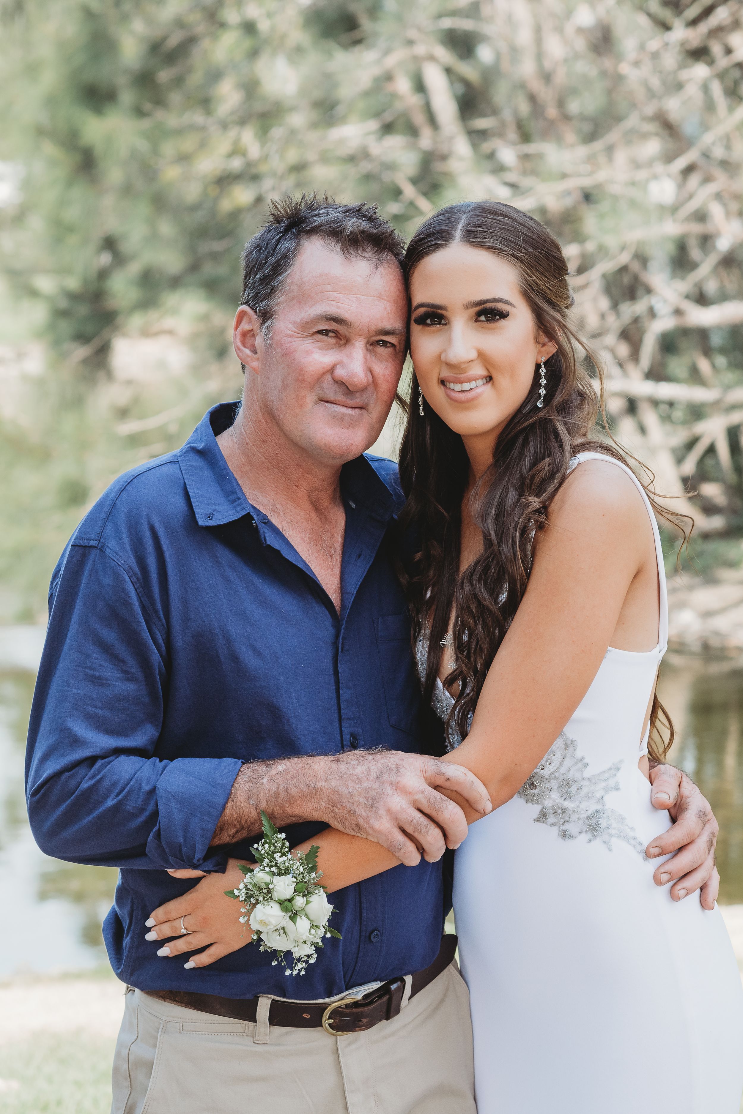 Wedding day portrait with bride in white dress and corsage alongside family member in blue shirt.