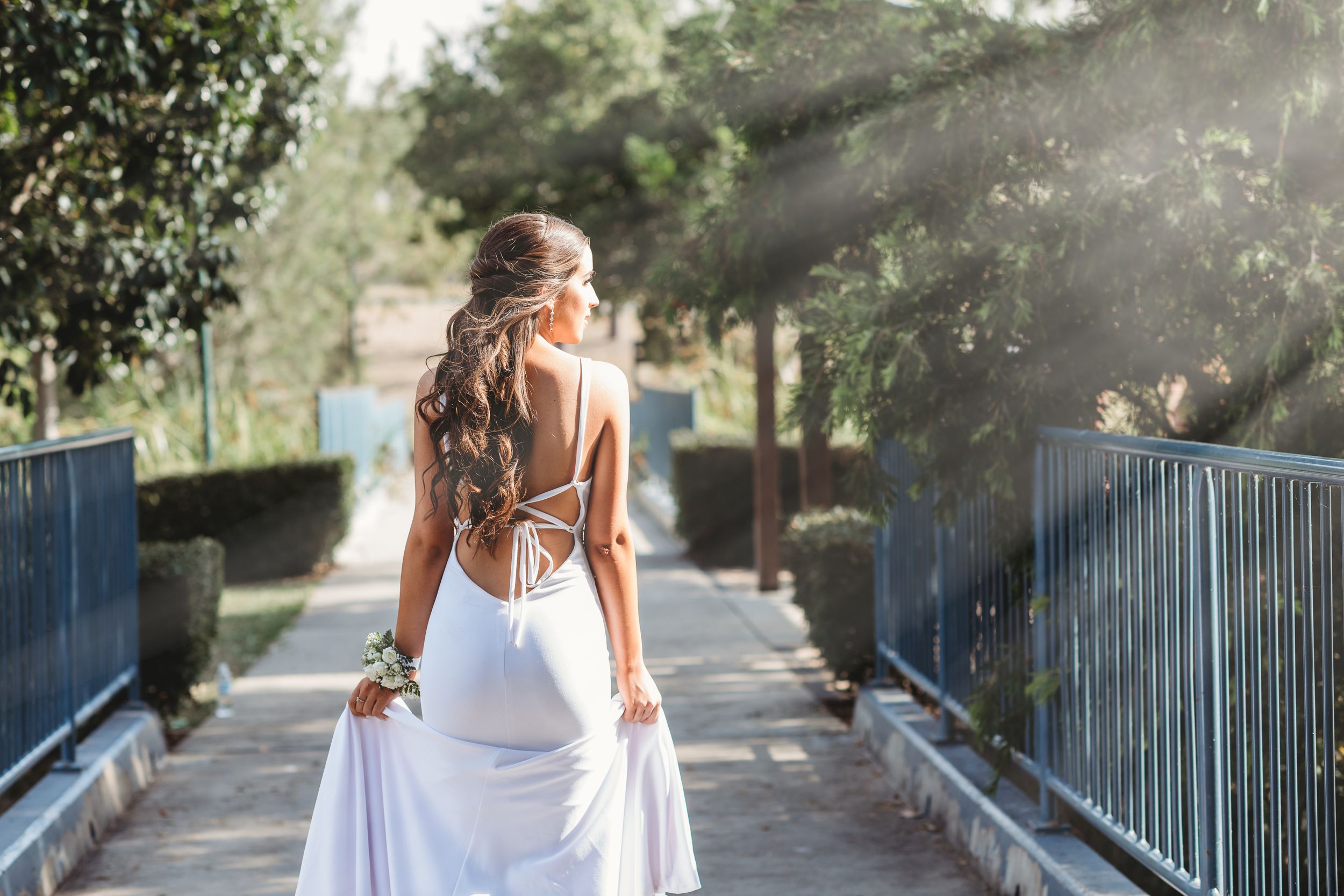 Bride in backless white wedding gown standing on bridge with sunlight streaming through trees.