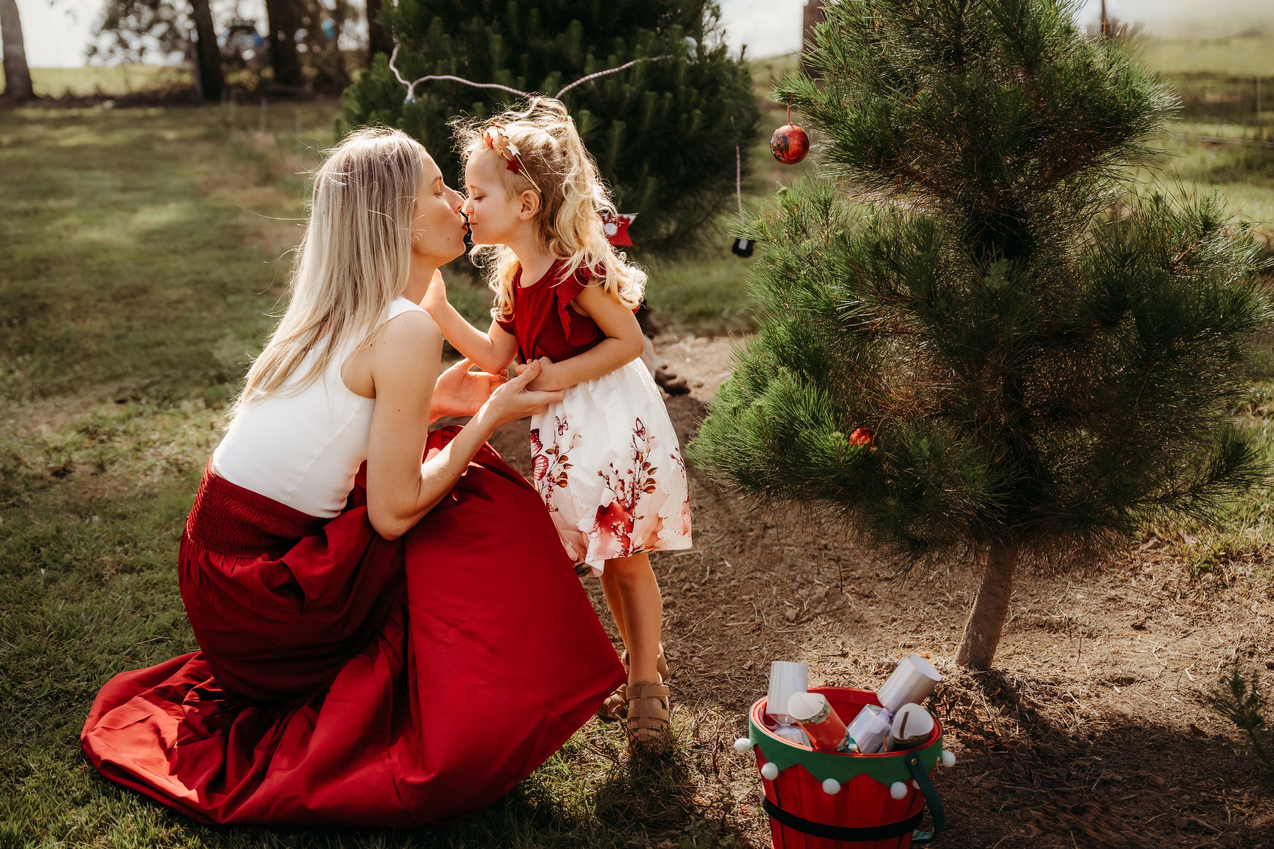 Two people in red dresses sit beside a small Christmas tree with wrapped presents beneath it in a garden setting.
