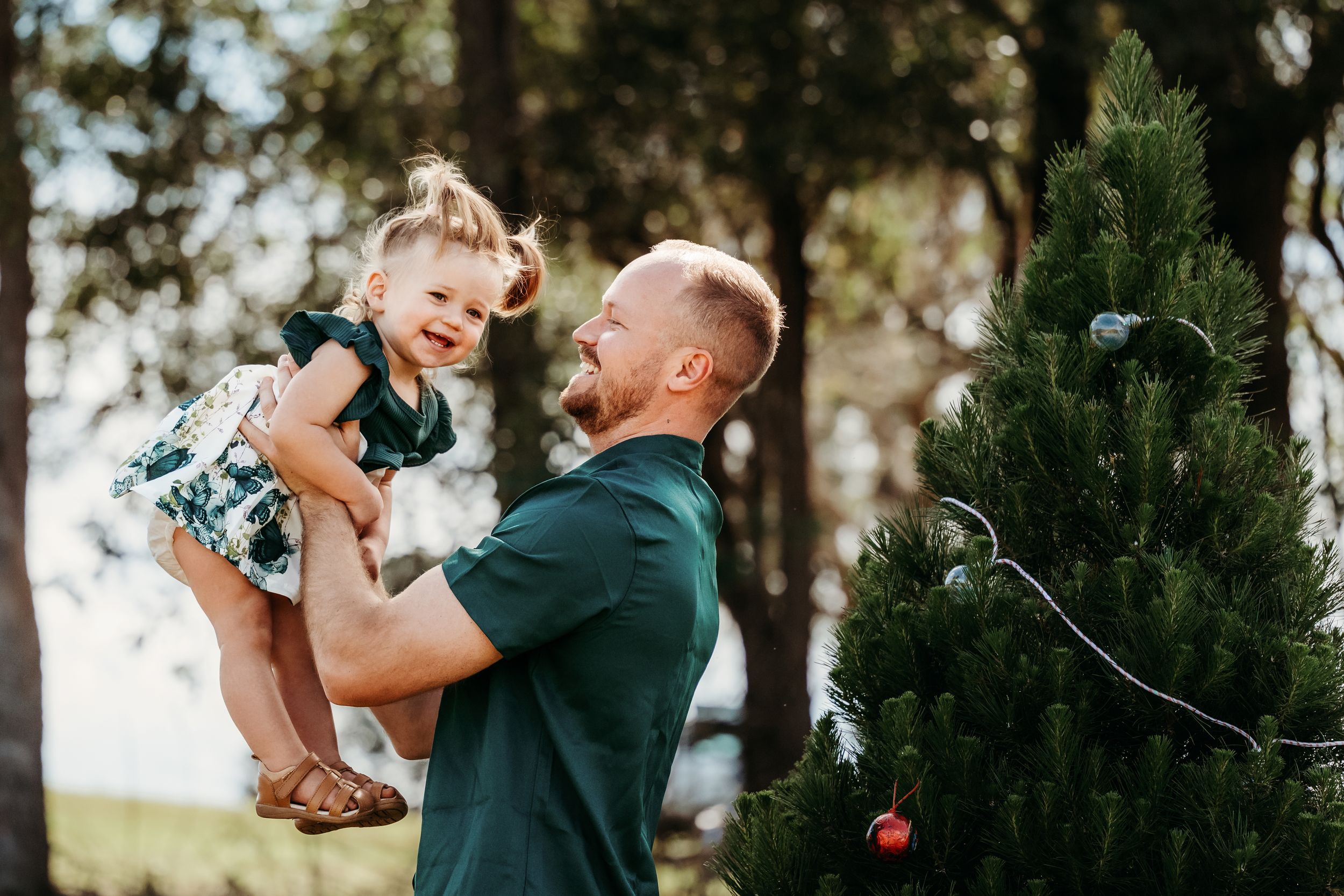 A happy couple holding their small dog while standing next to an evergreen tree in an outdoor setting.