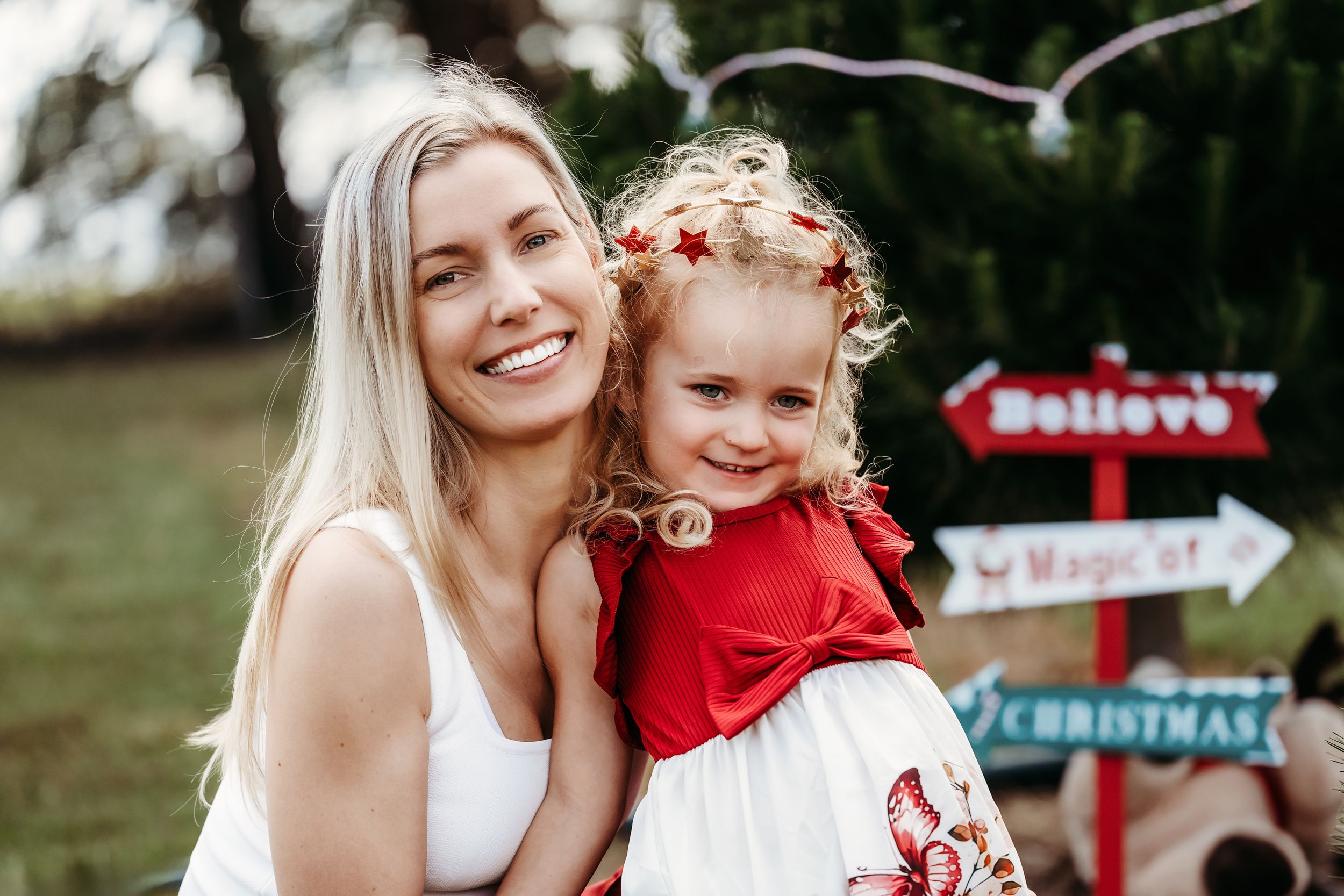 Parent and child pose outdoors by festive Christmas directional signs while wearing coordinated red and white outfits.
