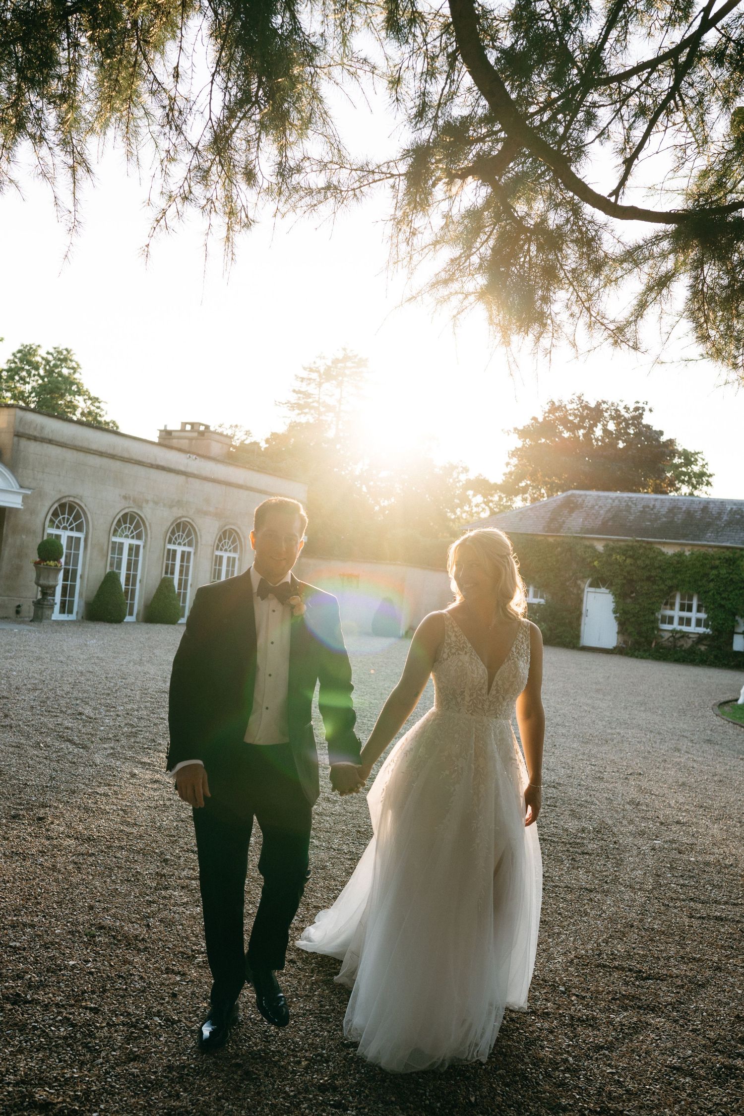 A couple walks hand in hand on a gravel path outside an elegant venue during golden hour sunset.