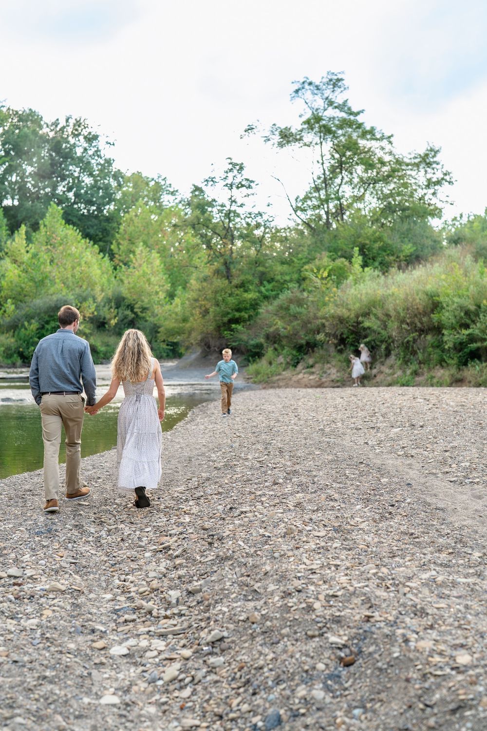 A family walks together on a rocky riverbank surrounded by lush green trees on a sunny day in Cleveland, Ohio for a Family Photo Session