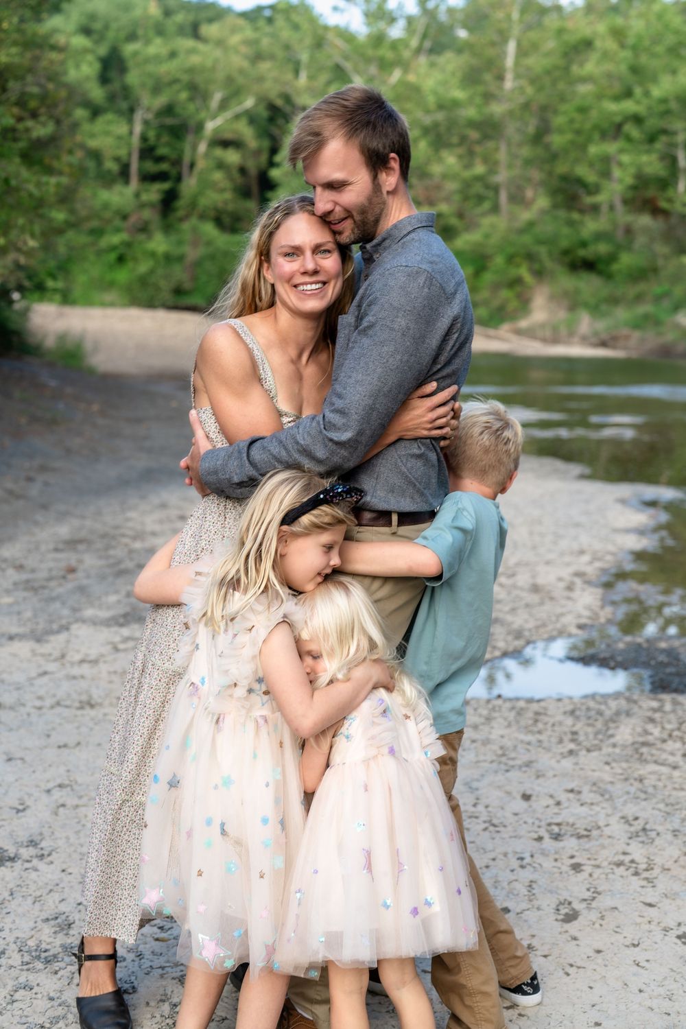 A family shares an intimate embrace while standing by a scenic river on a summer day at Brecksville Reservation Family Photo Session in CLE