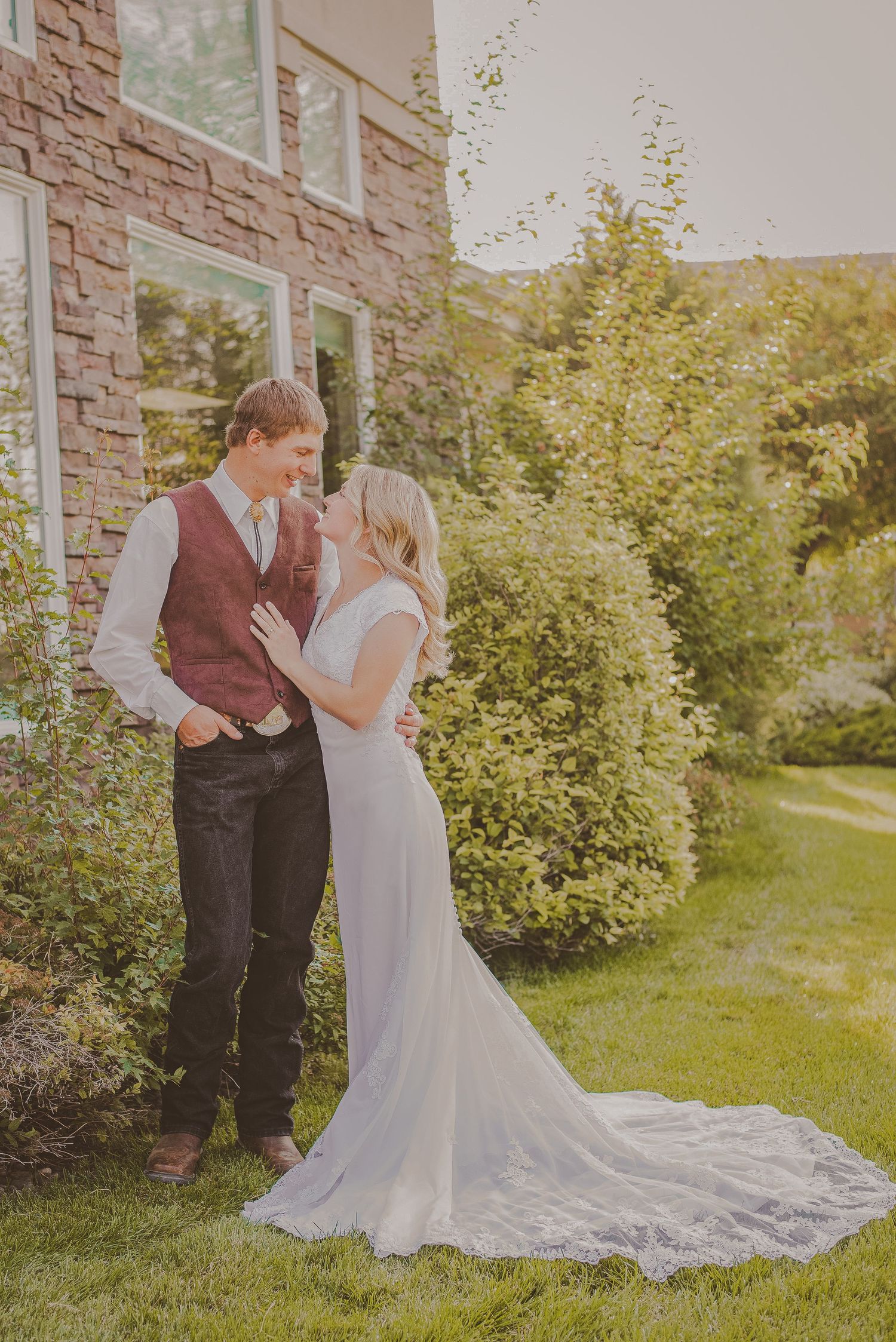 A wedding couple poses together against a stone building exterior surrounded by lush greenery.