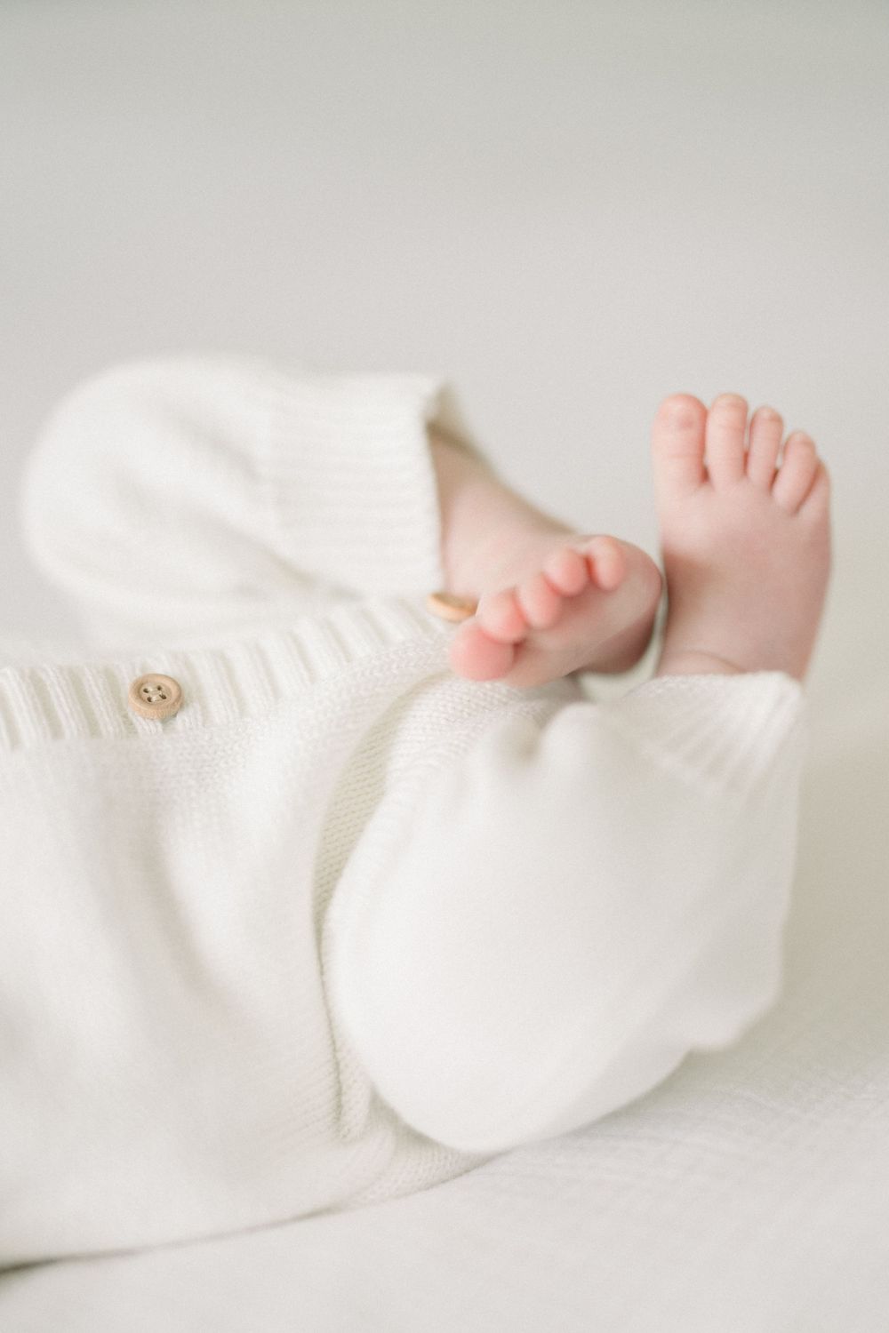 Tiny baby feet peek out from a soft white knitted sweater against a light background.