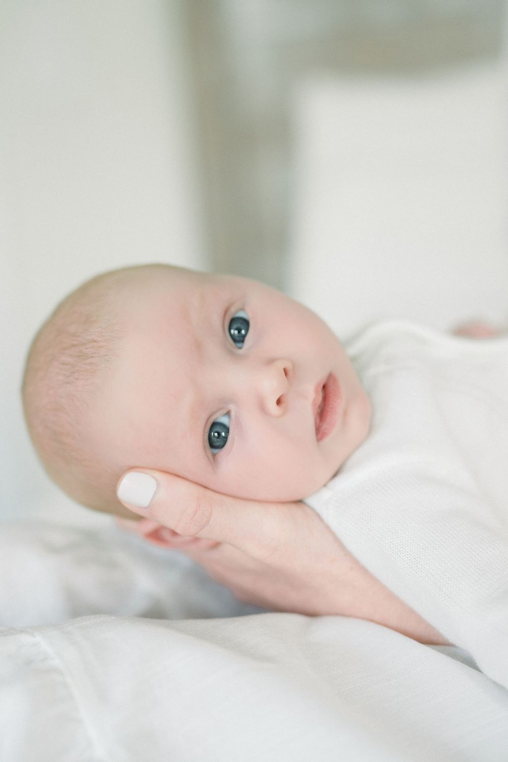 A sweet newborn baby with bright blue eyes lying on white bedding looking at the camera.