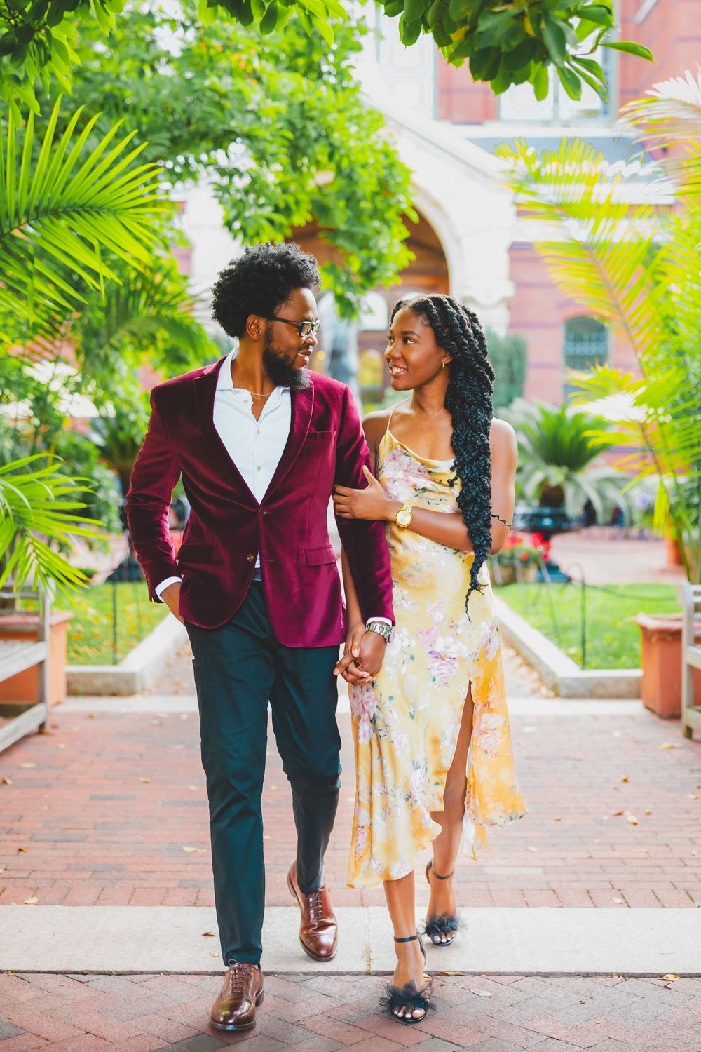 A stylish couple walks hand in hand along a brick pathway surrounded by tropical plants and palm fronds.
