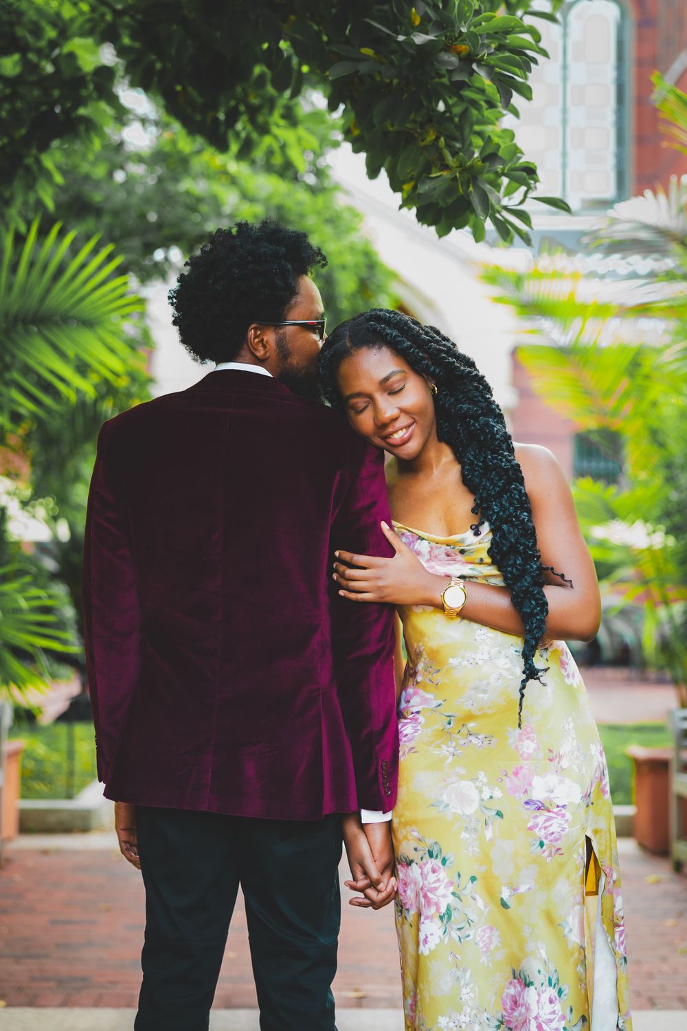 A couple shares romantic moments in an outdoor garden setting wearing burgundy and floral yellow outfits.