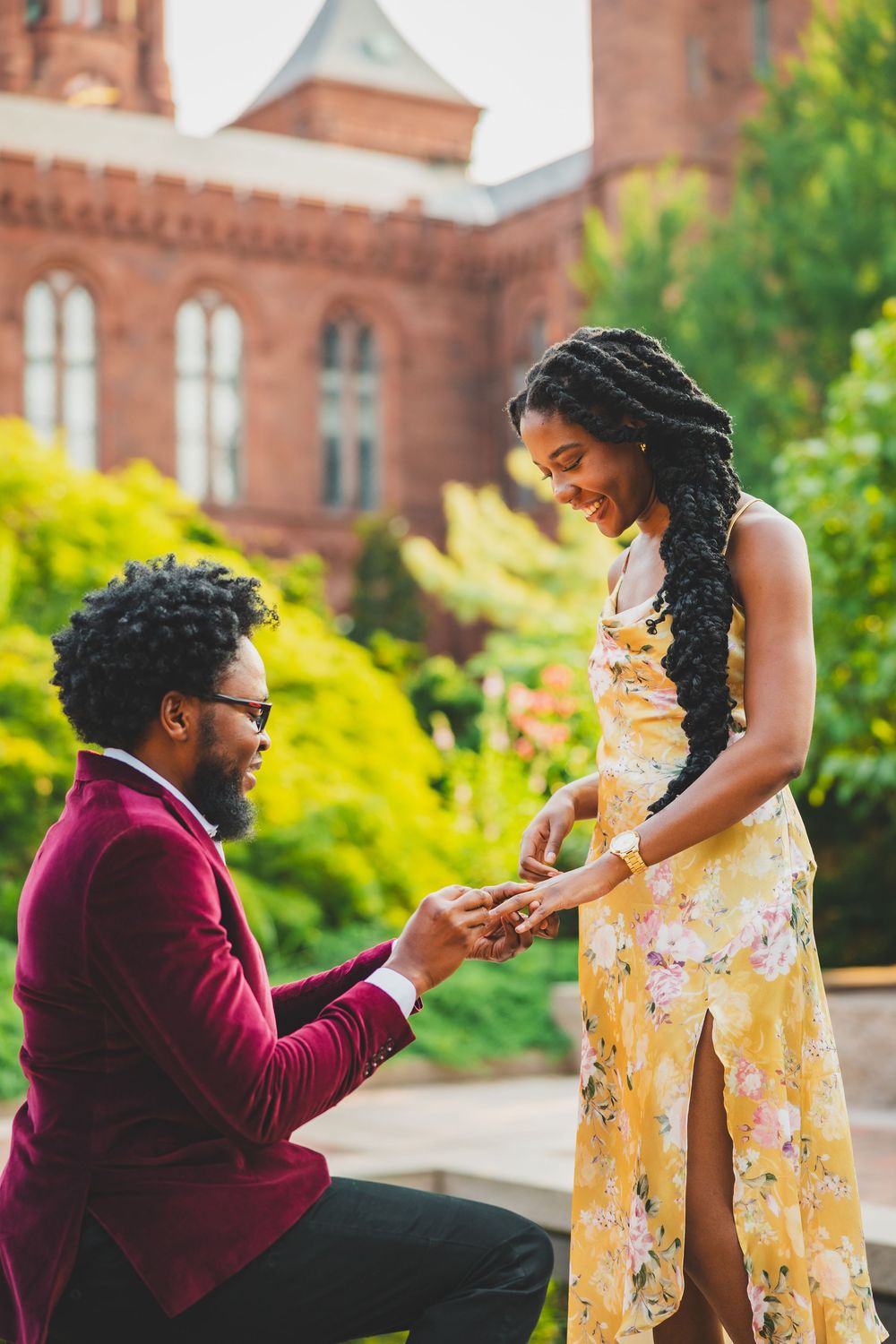 A romantic marriage proposal scene outdoors on a sunny day with brick buildings and green foliage in the background.