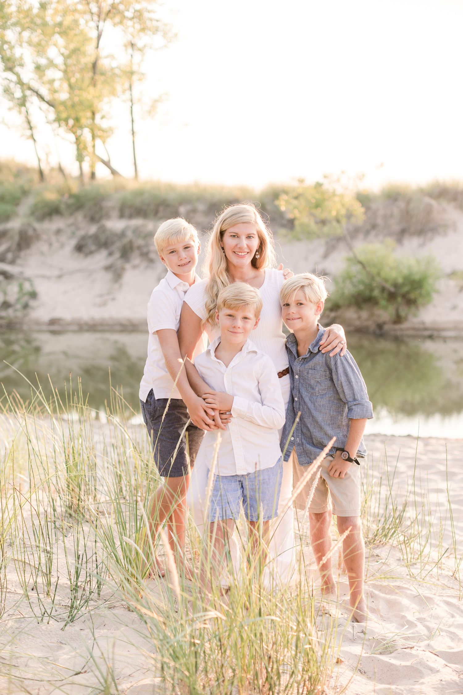 Children play and laugh together on a sandy beach near a lake during golden hour with tall grasses in the foreground.
