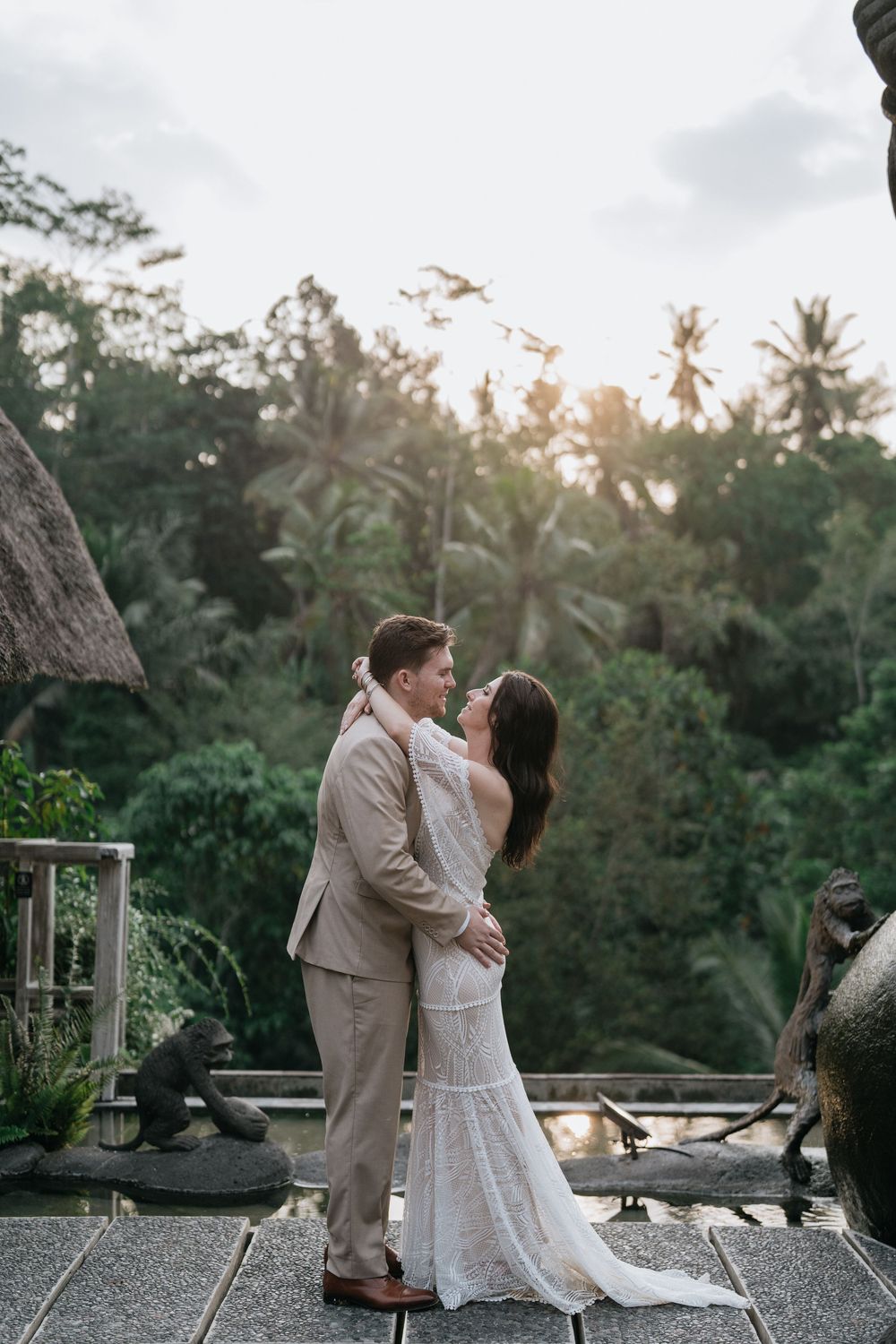 A romantic wedding couple shares a tender moment on a terrace surrounded by lush tropical greenery at sunset.