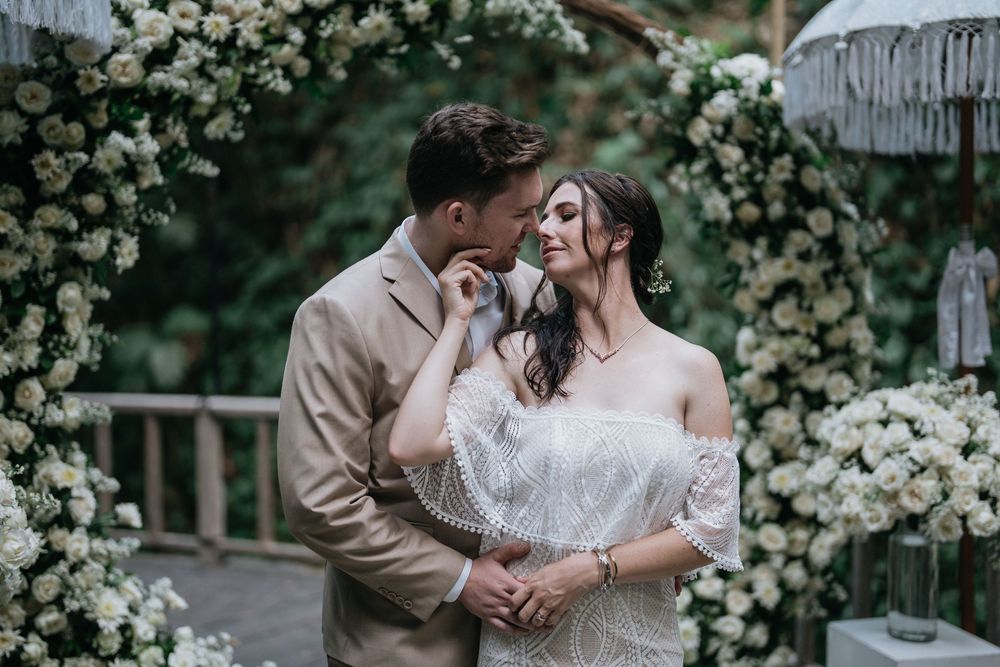 Wedding couple poses near a beautiful white floral arch in a garden setting.