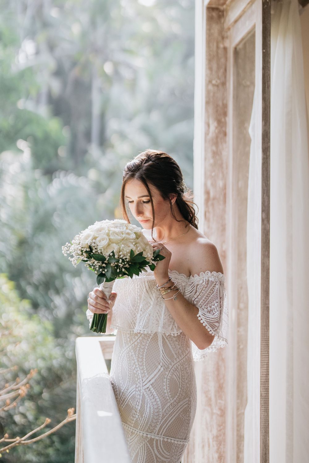 Bride in detailed lace gown holds a white bouquet near sunlit windows.