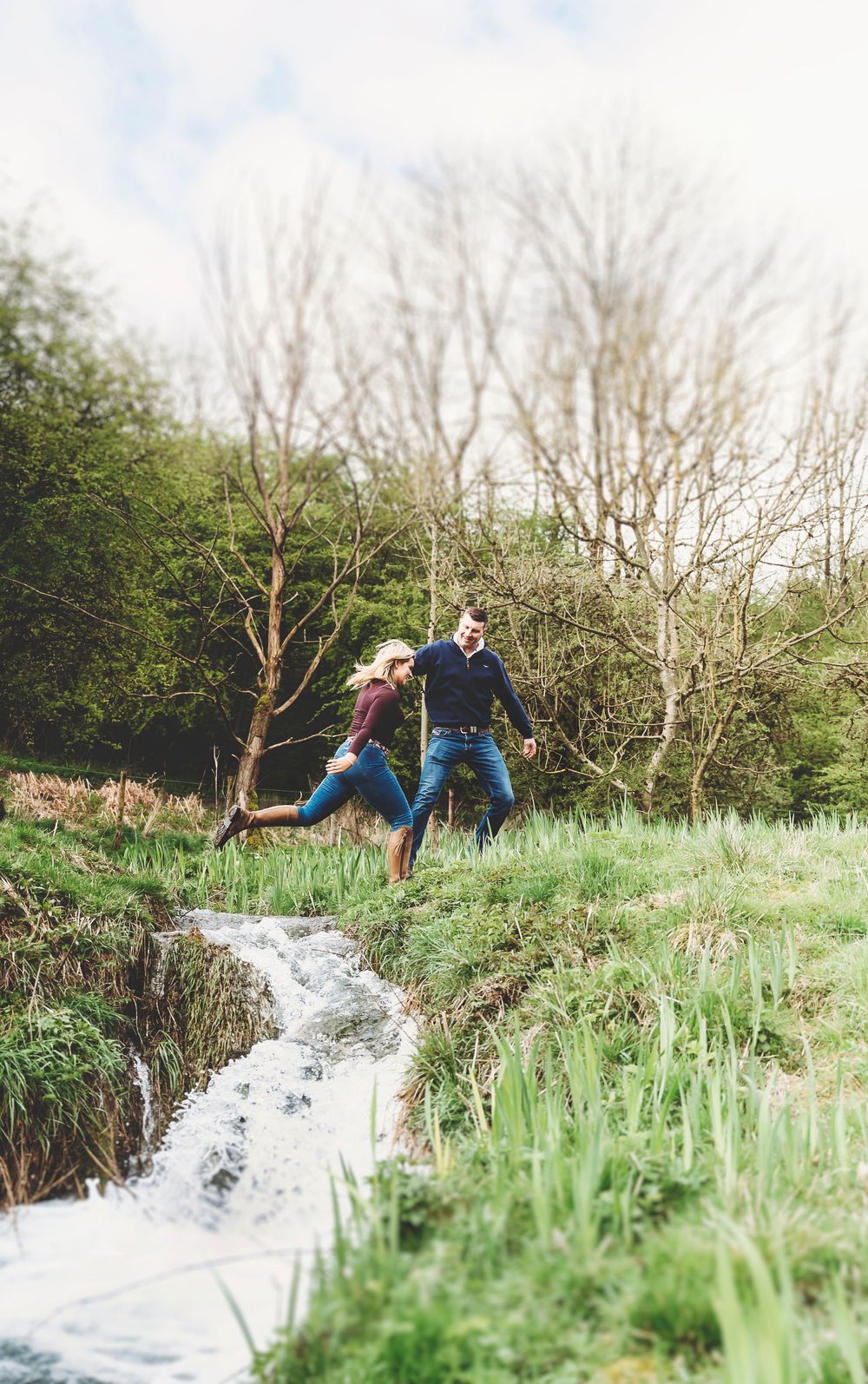 A person in blue jeans leaps across a rushing stream in a lush green landscape with bare trees in the background.