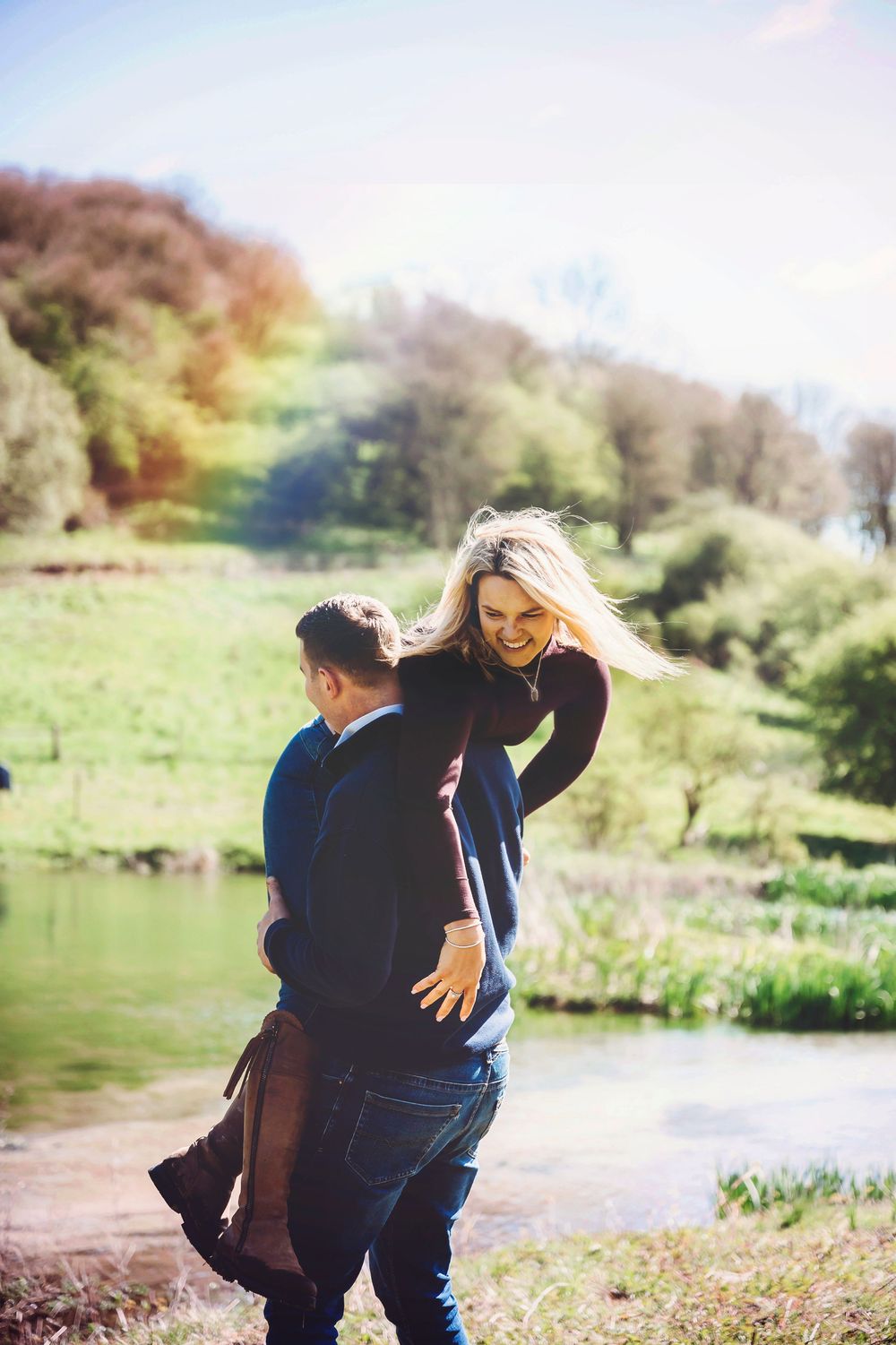 A couple embraces playfully by a serene lake surrounded by autumn trees and green grass.