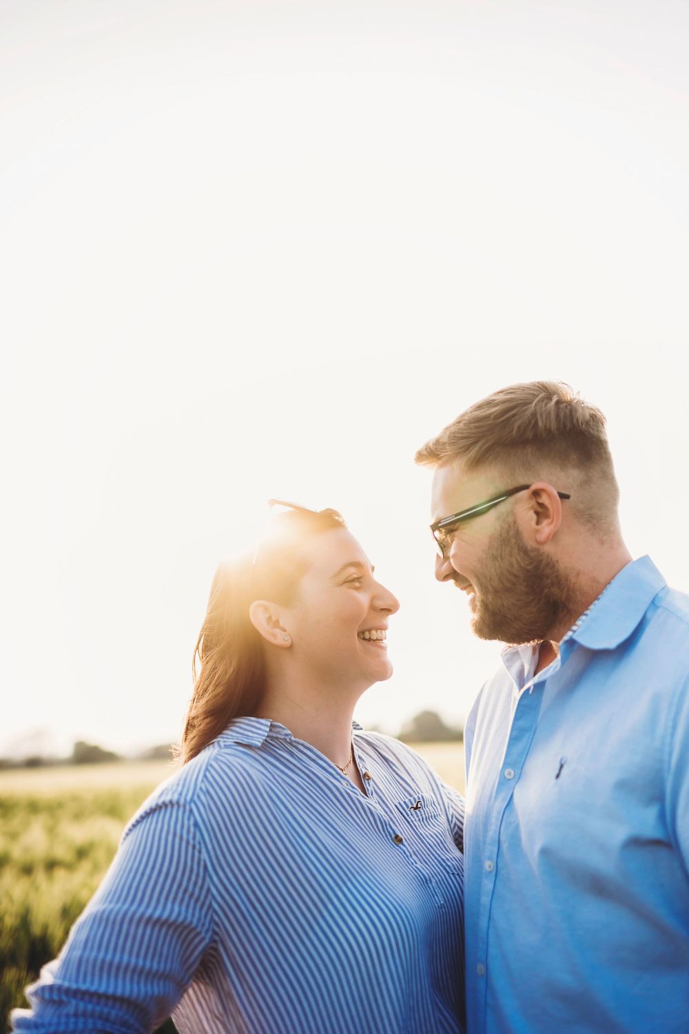 Couple in blue shirts share tender moments during outdoor photoshoot at sunset in a field.