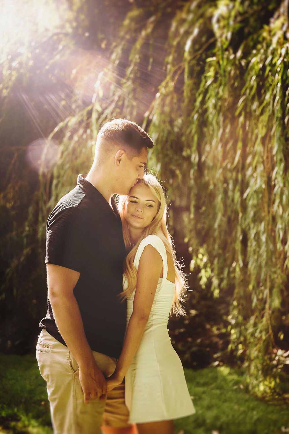 A couple shares a romantic moment under sunlit willow trees in a dreamy garden setting.
