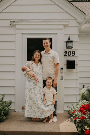 A family sits together on the front steps of a white sided house with red flowers in the garden at 209.