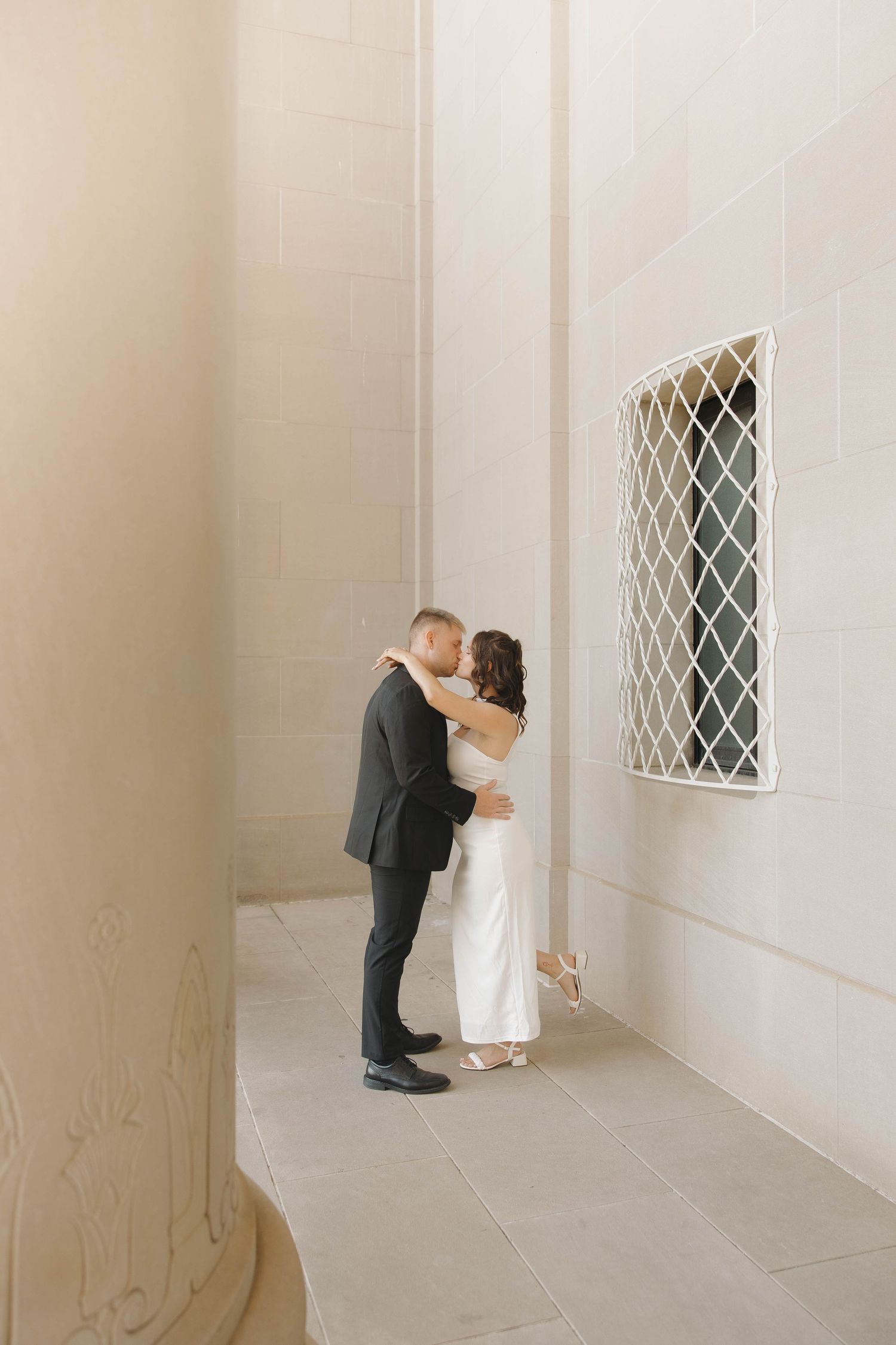 A couple shares a kiss next to an architectural window detail in a minimalist white hallway setting.