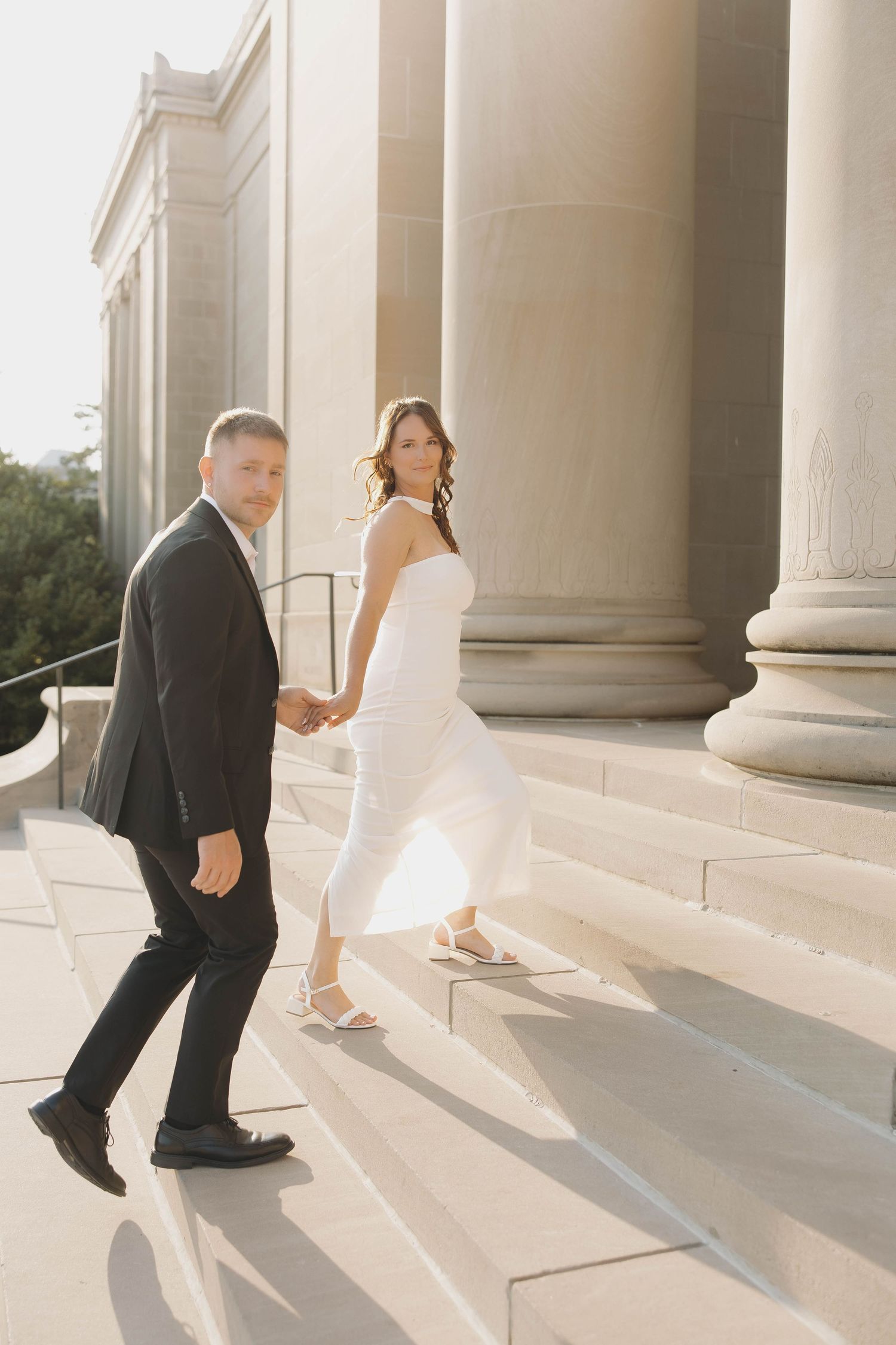Newlywed couple walking up elegant stone steps in sunlight, showing bride's white dress and groom's black suit details.
