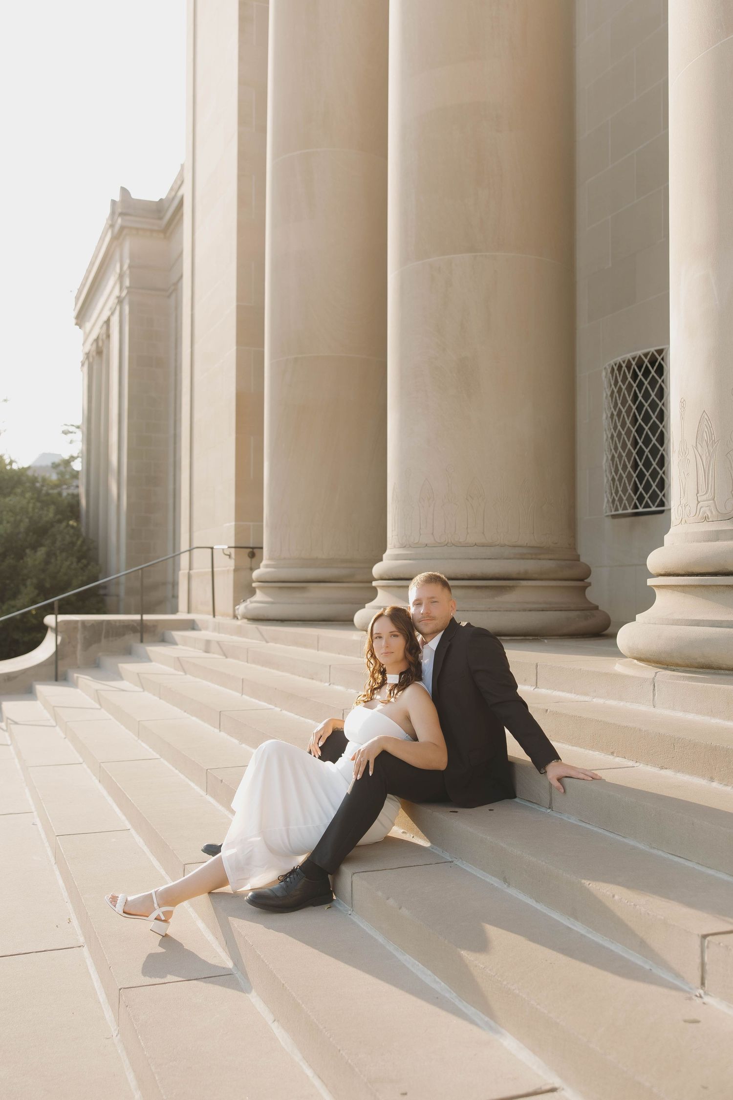 A couple in formal wedding attire sits intimately on elegant white stone steps between grand pillars in sunlit setting.