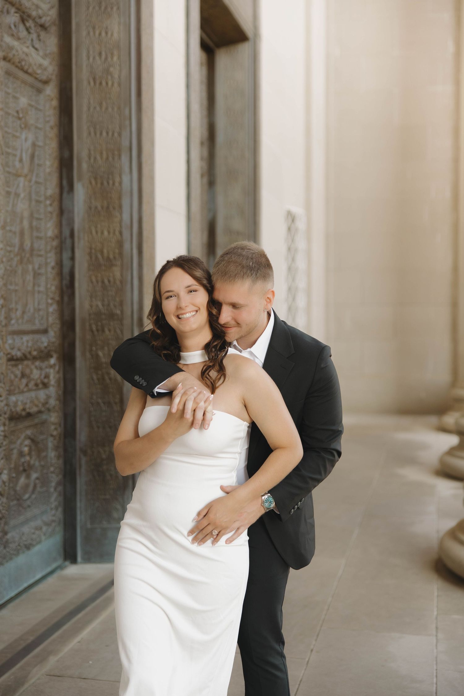 A romantic wedding couple embraces and shares a playful moment in an elegant stone colonnade hallway.