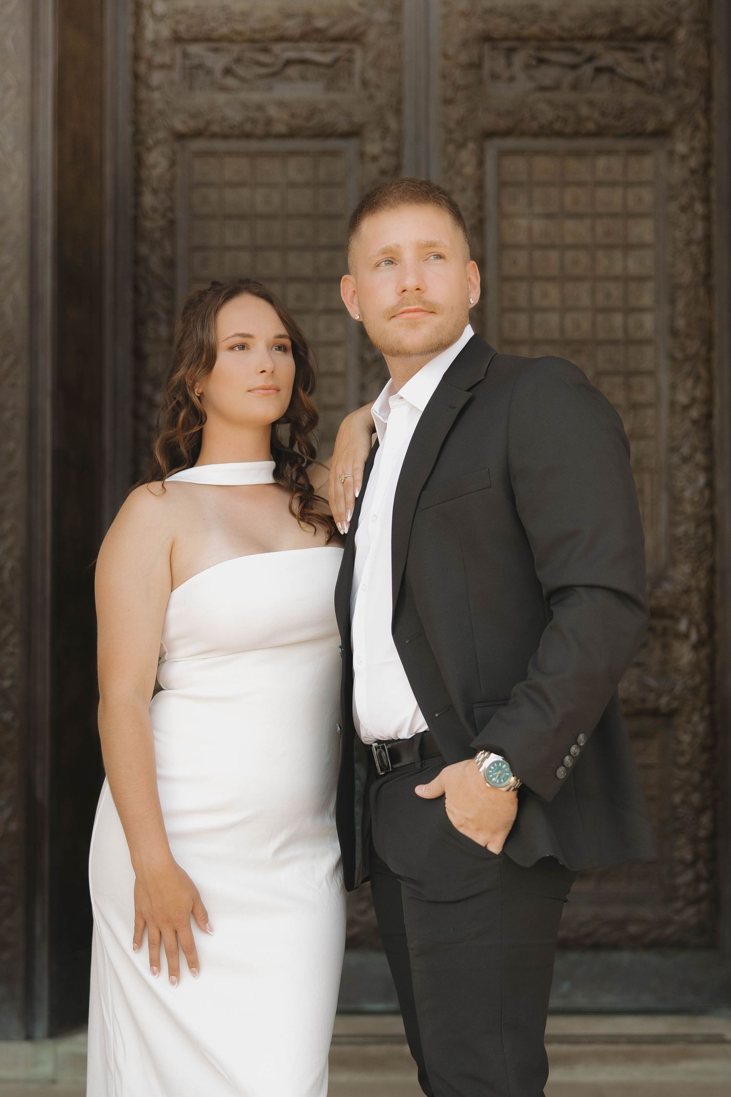 Wedding couple poses against ornate stone wall backdrop in formal attire with dramatic lighting.