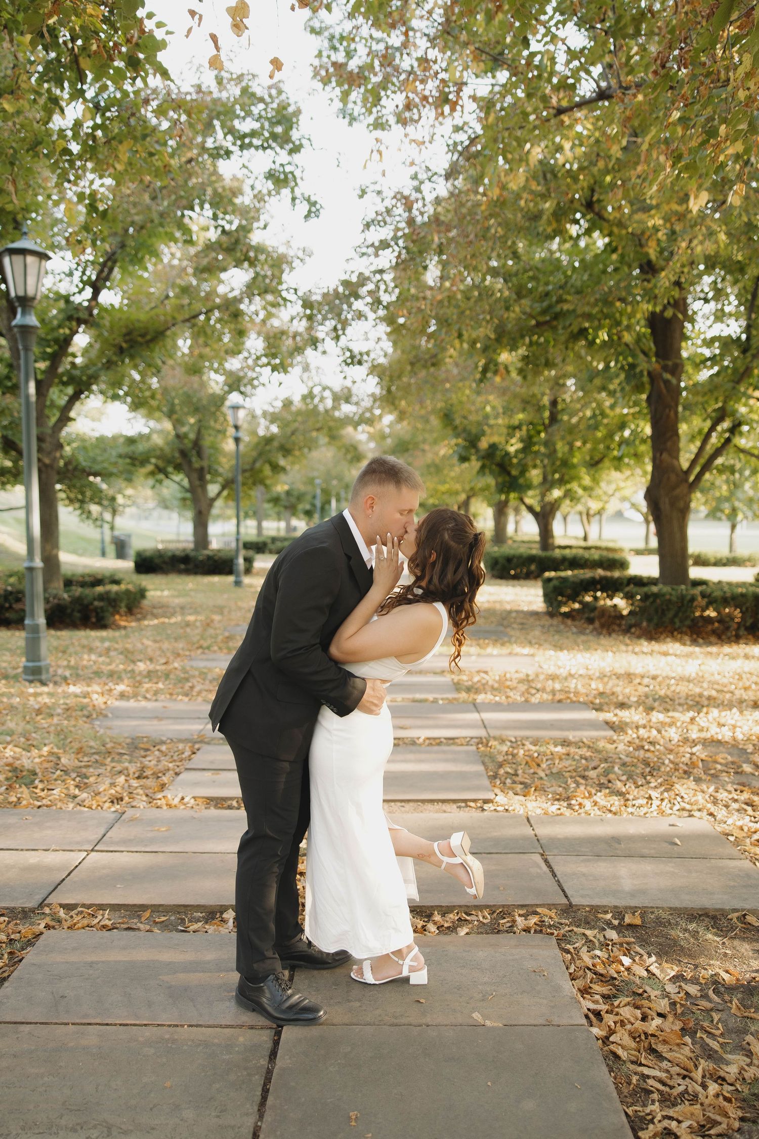 A couple sits together on a sidewalk surrounded by fall leaves while leaning in to kiss.