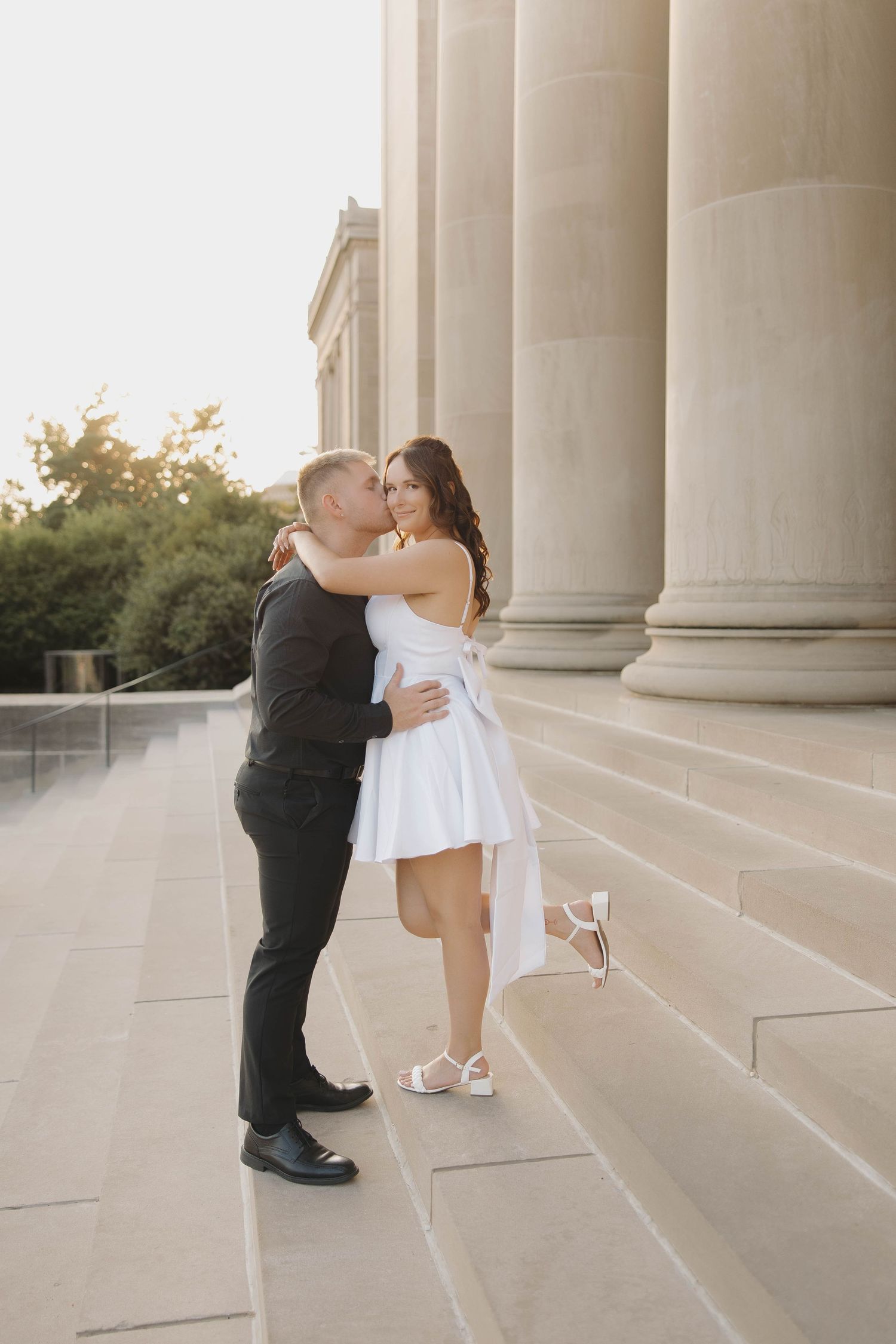 A couple in formal attire shares a romantic moment between grand columns at sunset during their wedding photoshoot.