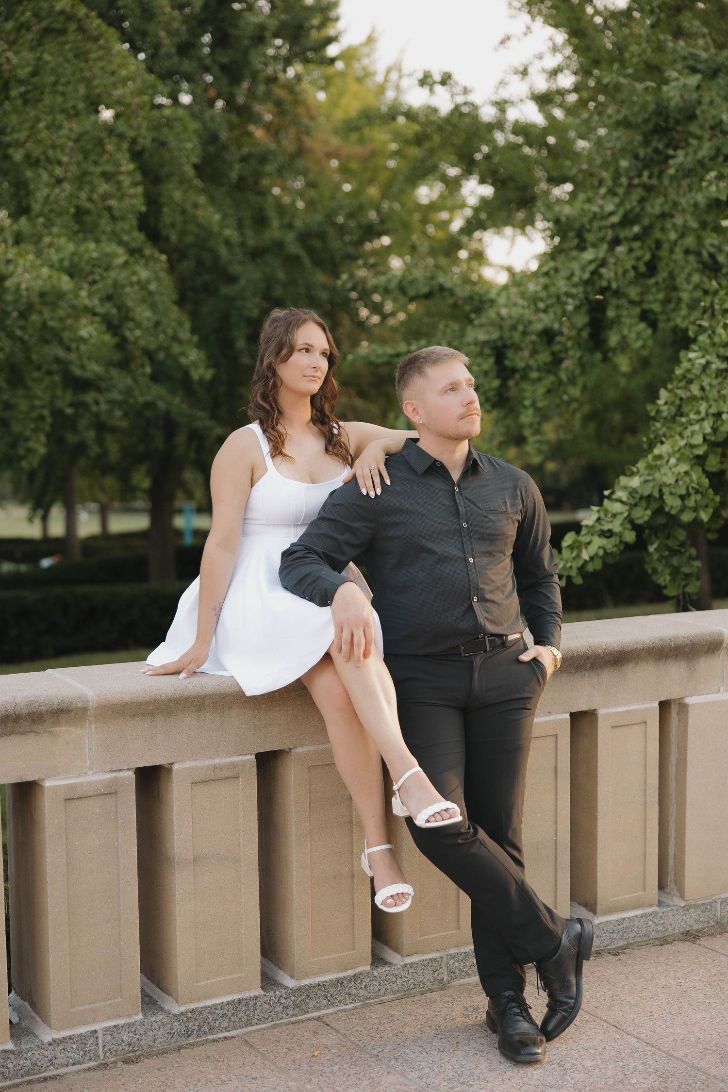 An engaged couple poses casually along a stone balcony surrounded by green trees during an outdoor photoshoot.
